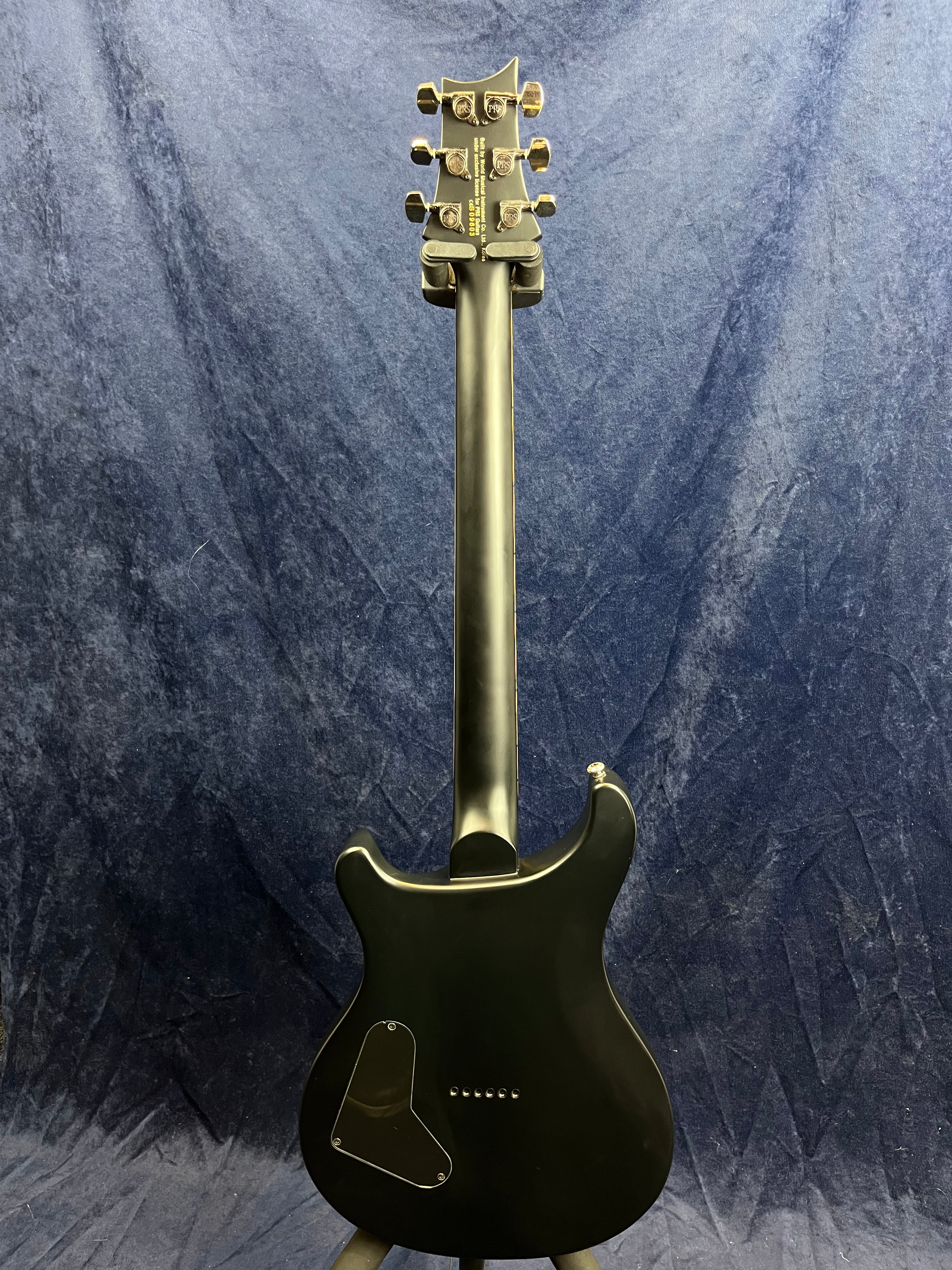 Matte black electric guitar shown from the back, gold tuning pegs, contoured body, triangular electronics cover on lower left.