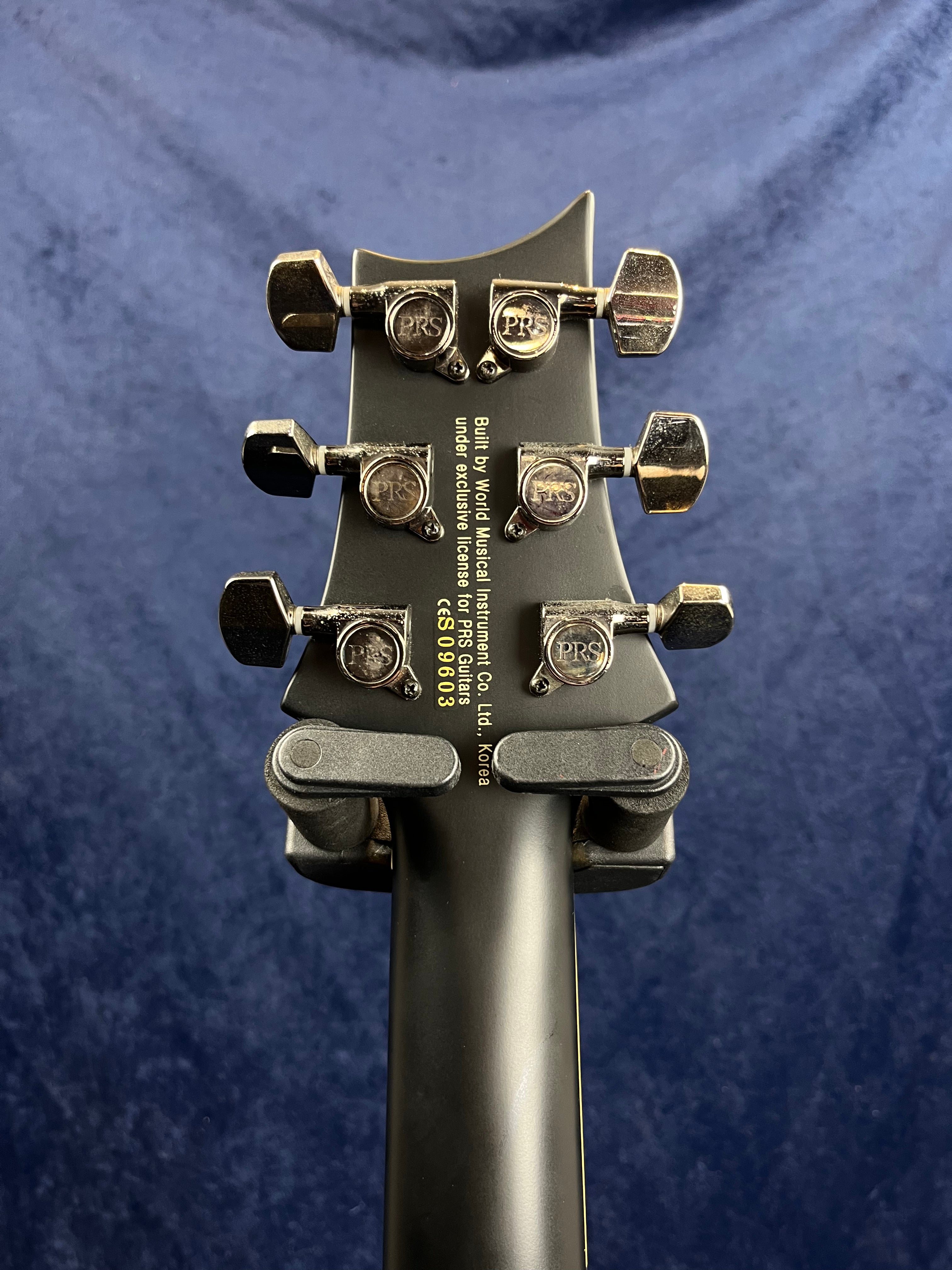 Six chrome PRS-branded tuning pegs on a matte black electric guitar headstock, manufacturer information and serial number printed in yellow.