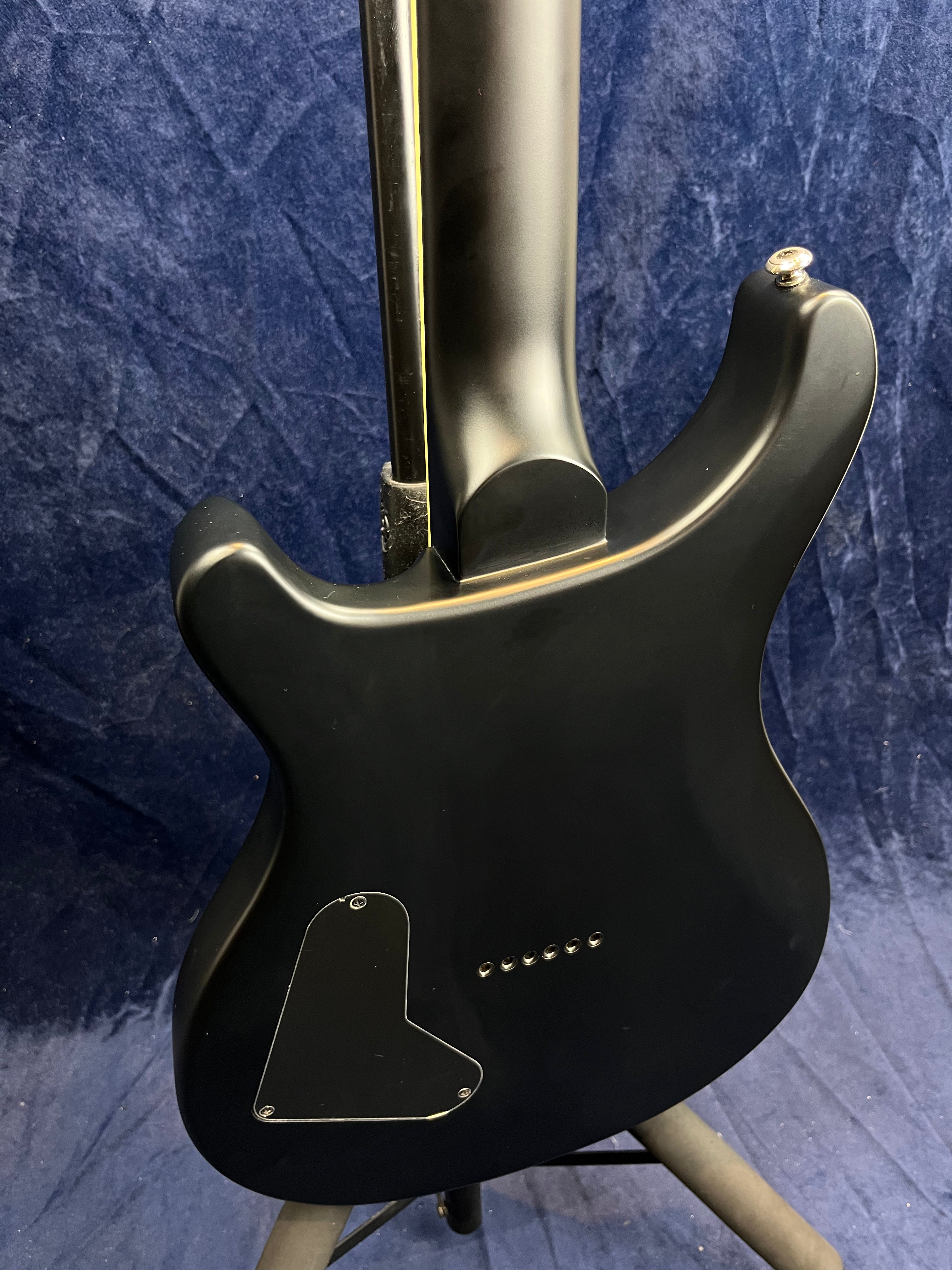 Matte black electric guitar back, showing a contoured neck joint, rear electronics cover, string ferrules, and strap button against a blue background.