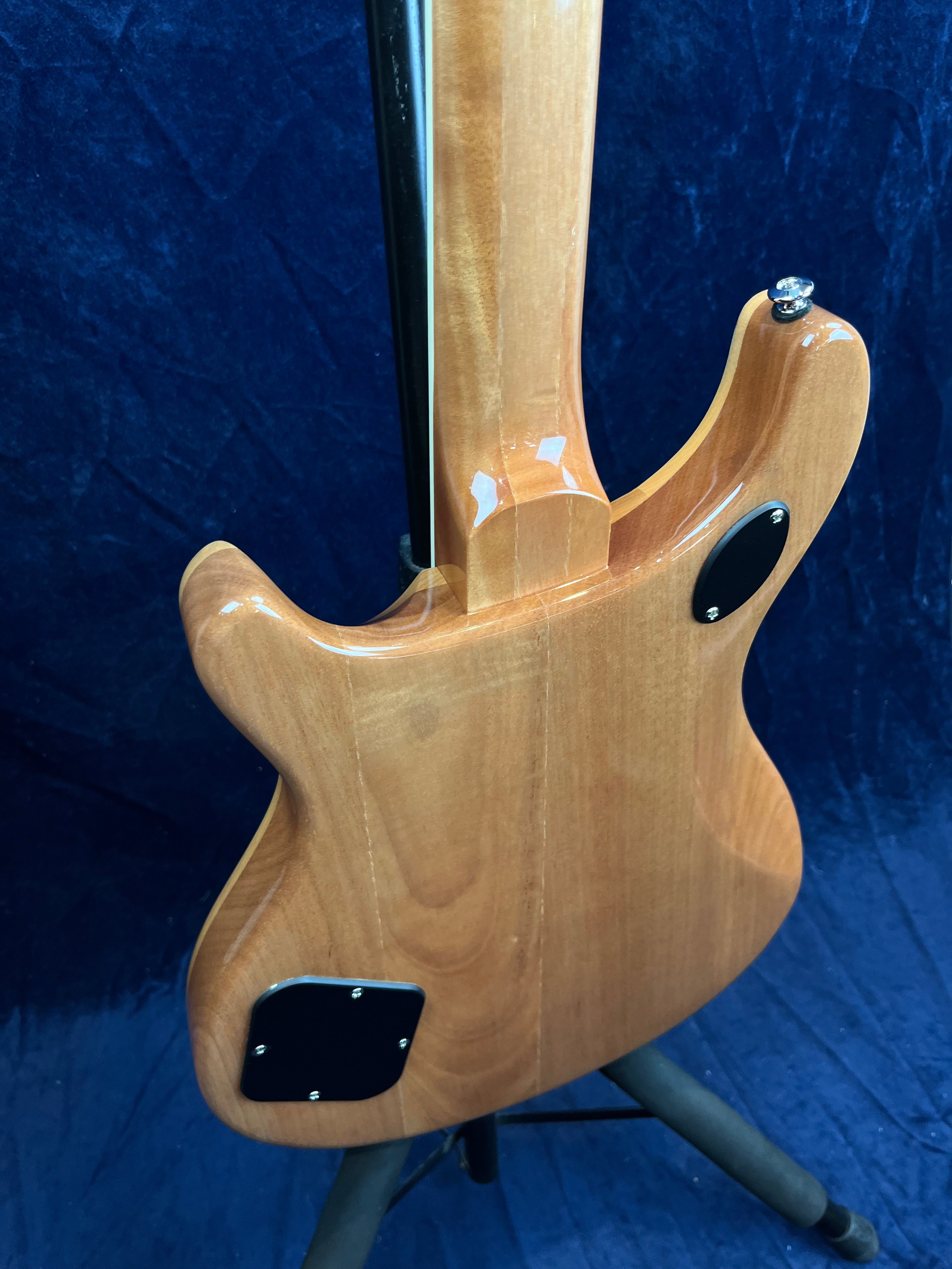Corps en bois verni d’une guitare électrique vu de dos, avec plaques noires vissées et manche assemblé, posé sur un support devant un fond bleu.