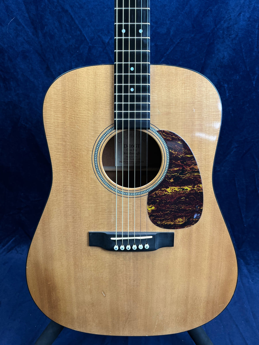 Natural wood acoustic guitar with a dark, tortoiseshell-pattern pickguard and six strings, set against a dark blue background.