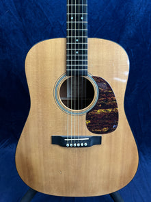 Natural wood acoustic guitar with a dark, tortoiseshell-pattern pickguard and six strings, set against a dark blue background.
