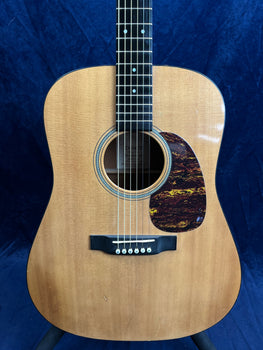 Natural wood acoustic guitar with a dark, tortoiseshell-pattern pickguard and six strings, set against a dark blue background.