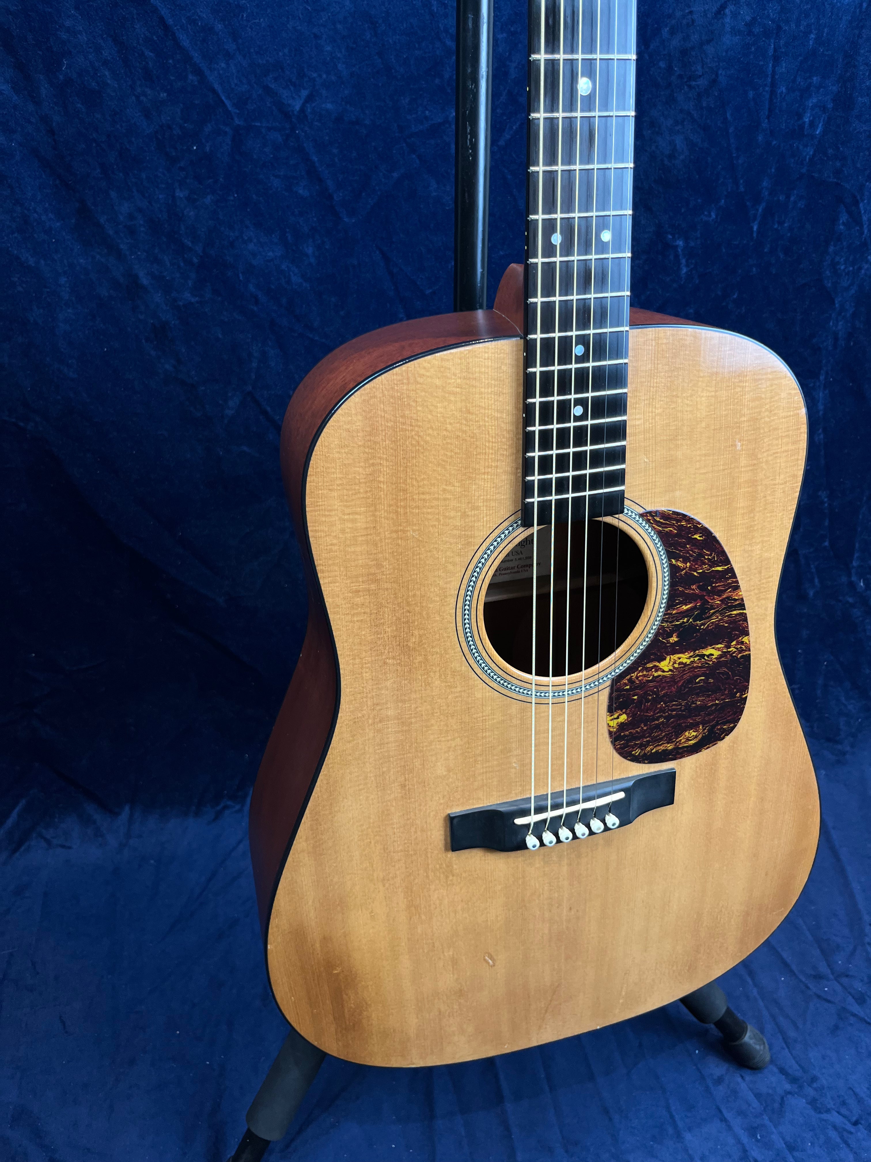 Wooden acoustic guitar with a natural finish, dark brown sides, six strings, and a tortoiseshell-style pickguard, shown on a stand.