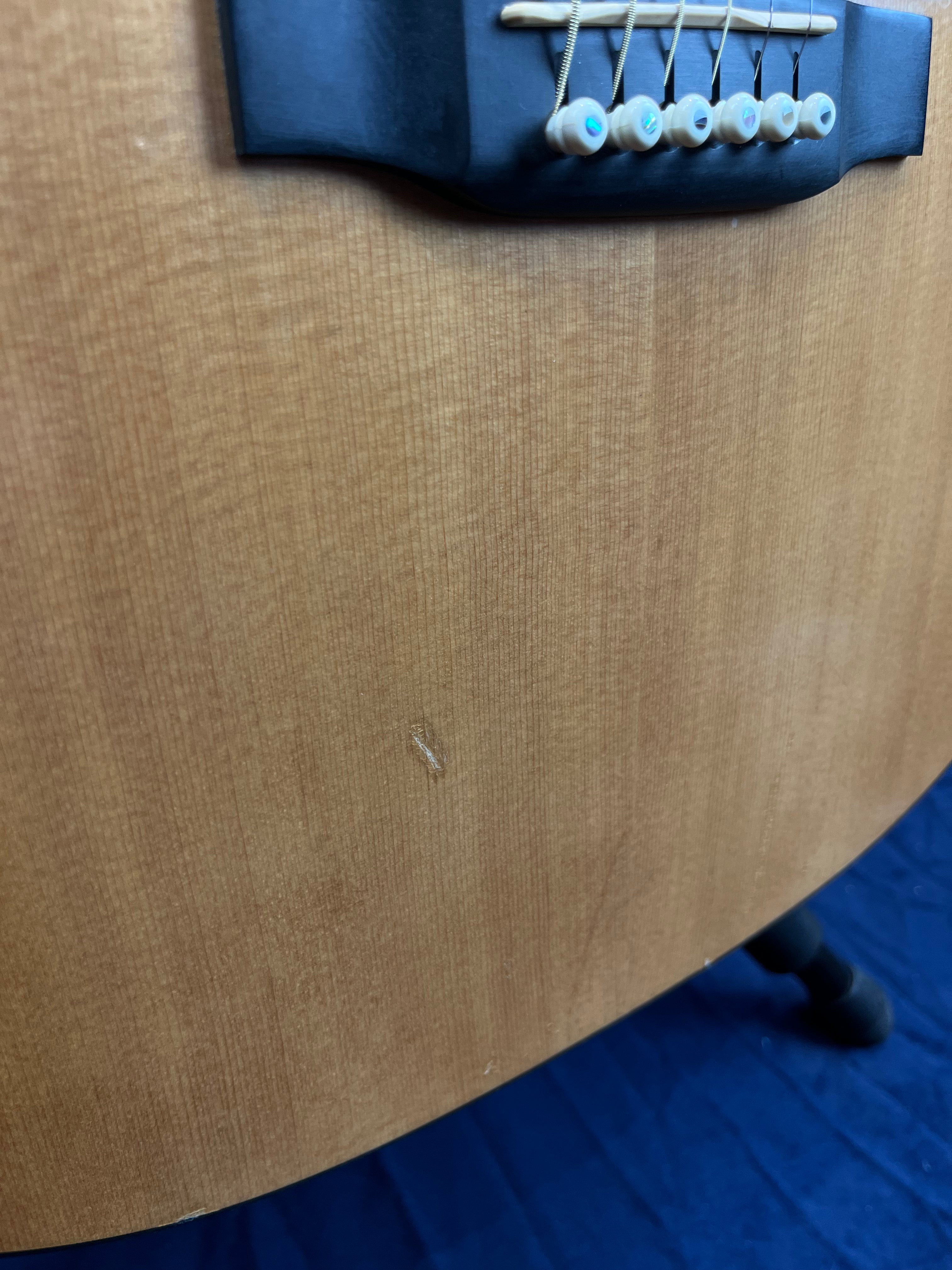 Lower body of an acoustic guitar showing a small scratch in the wood below the bridge and strings.
