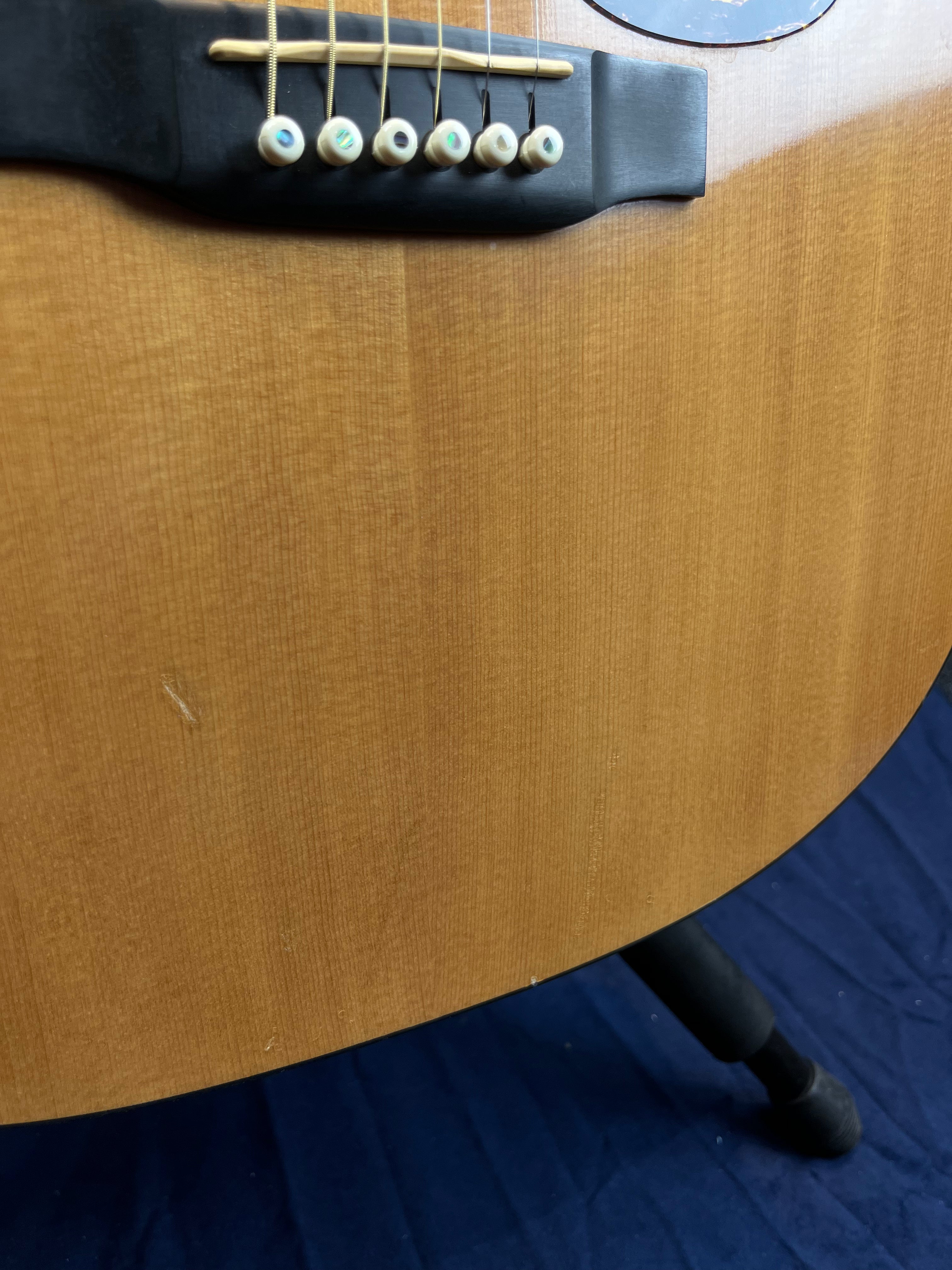 Close-up of an acoustic guitar’s wooden body, showing six bridge pins, strings, slight surface scratches, and part of a black guitar stand.