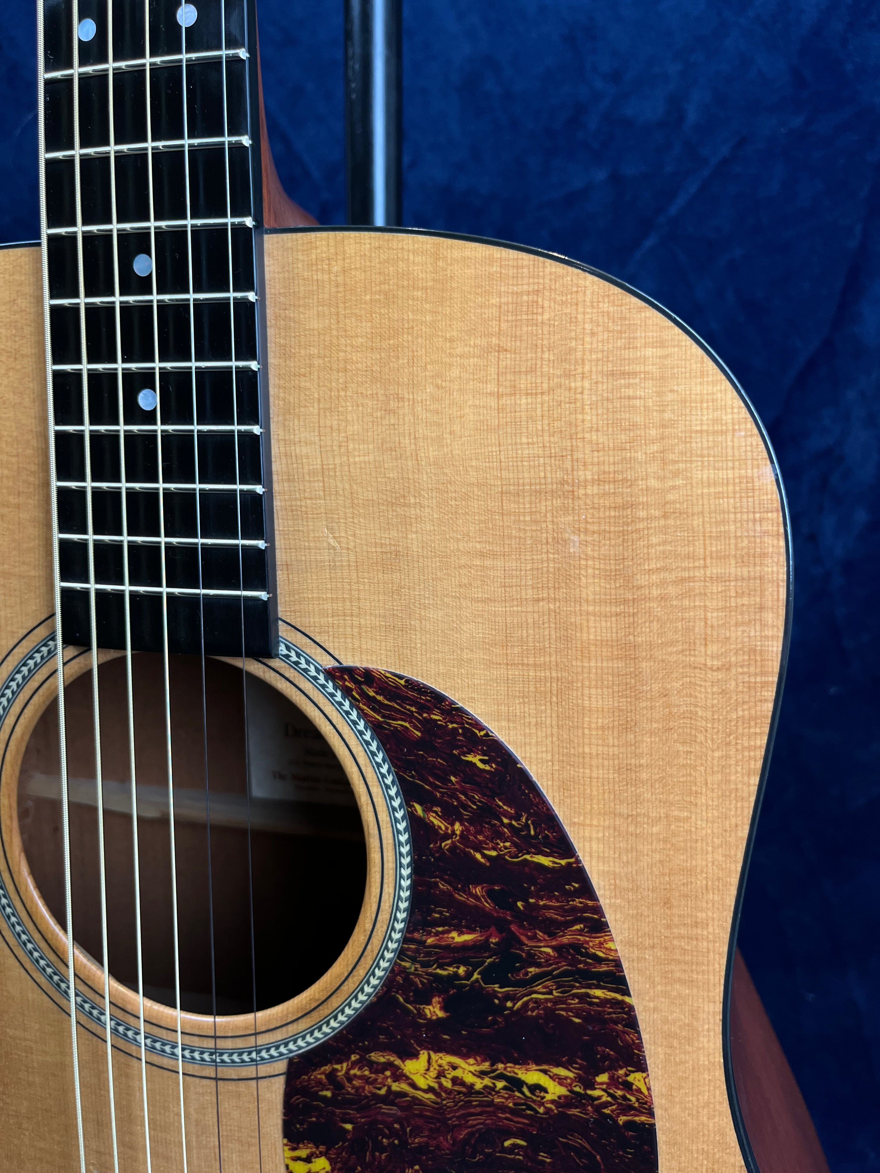 Light wood acoustic guitar body with dark tortoiseshell-pattern pickguard, black fretboard, and metal strings against a dark blue background.