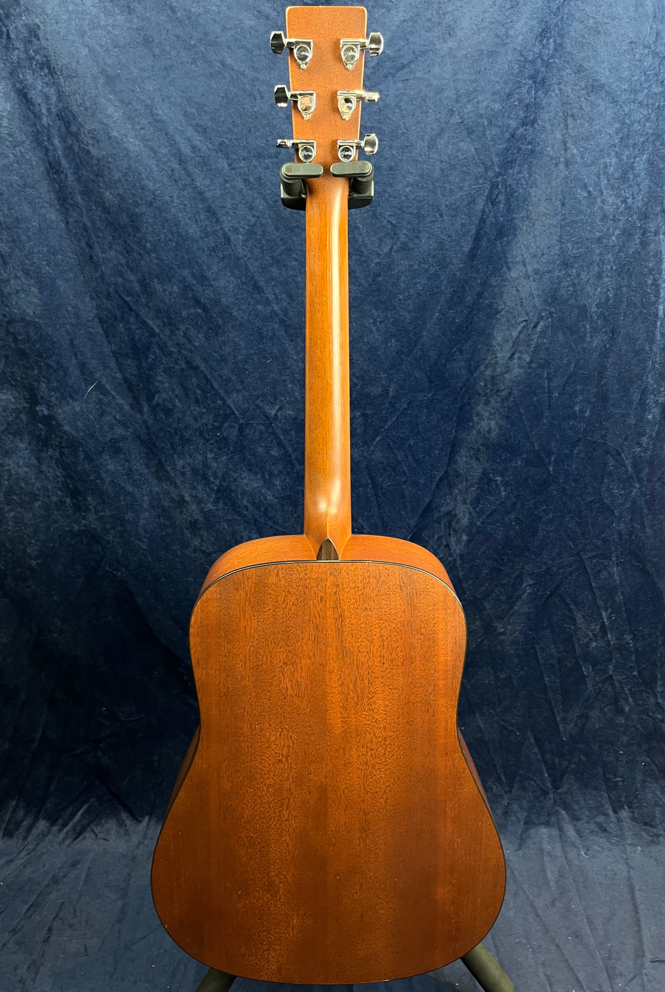 Mahogany acoustic guitar shown from the back, highlighting wood grain, neck, and chrome tuning pegs against a dark blue background.