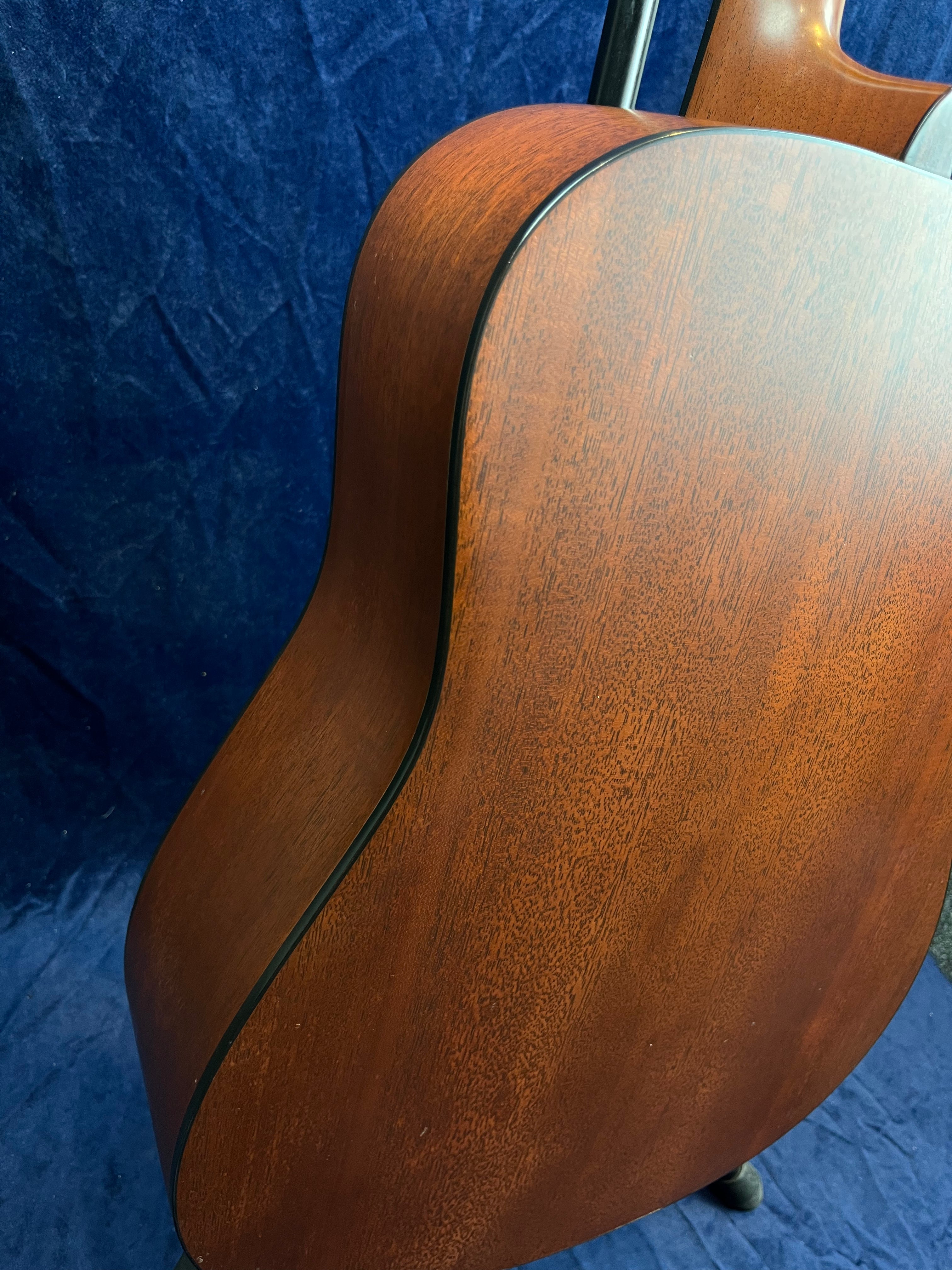 Mahogany-finished acoustic guitar body with smooth, matte wood grain and black binding, viewed from the back against a dark blue textured background.