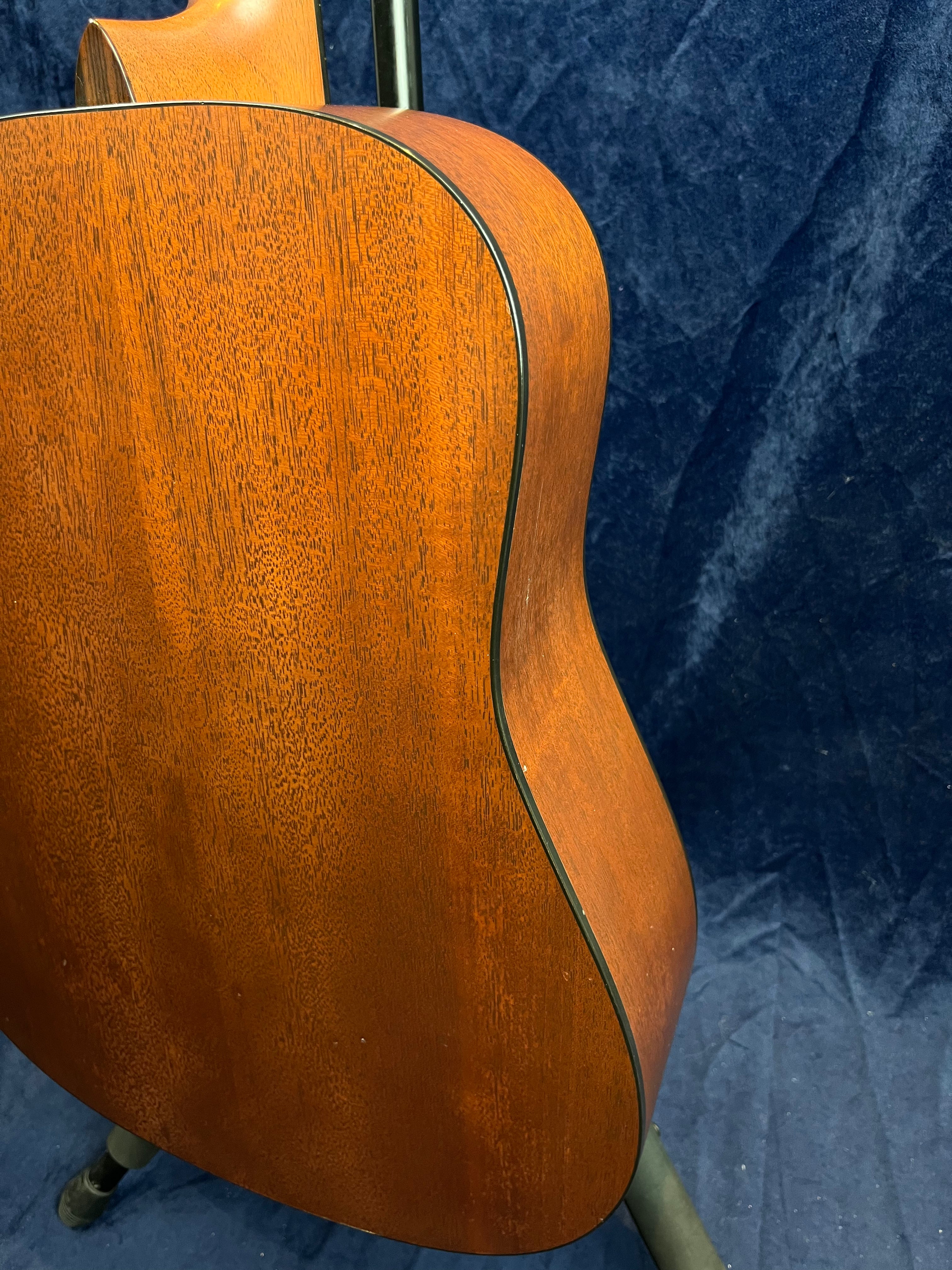 Back and side of an acoustic guitar with a reddish-brown wood grain finish, black edging, and a curved body resting on a stand against a dark backdrop.