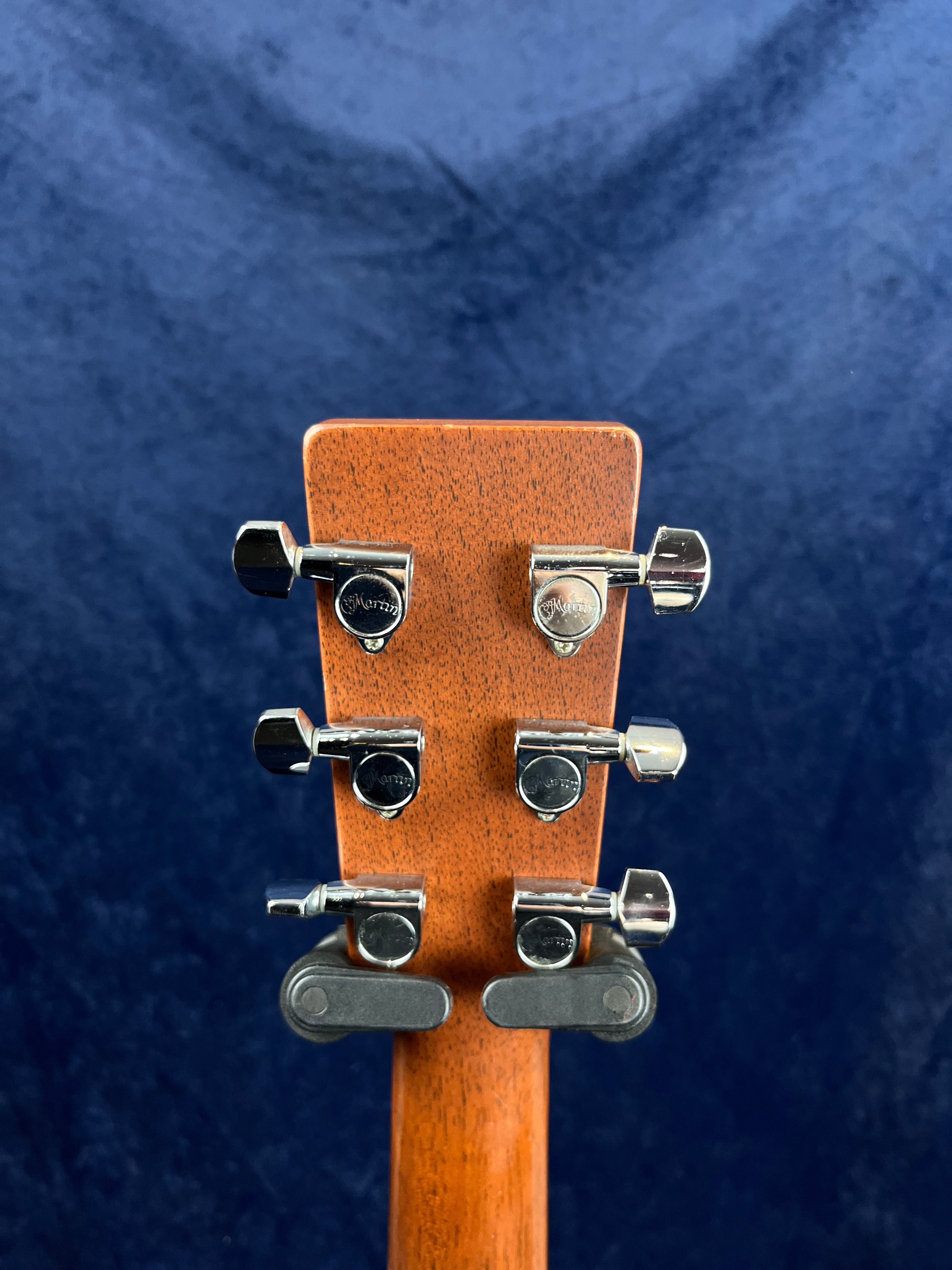 Brown acoustic guitar headstock with six chrome tuning pegs, viewed from the back against a dark blue background.
