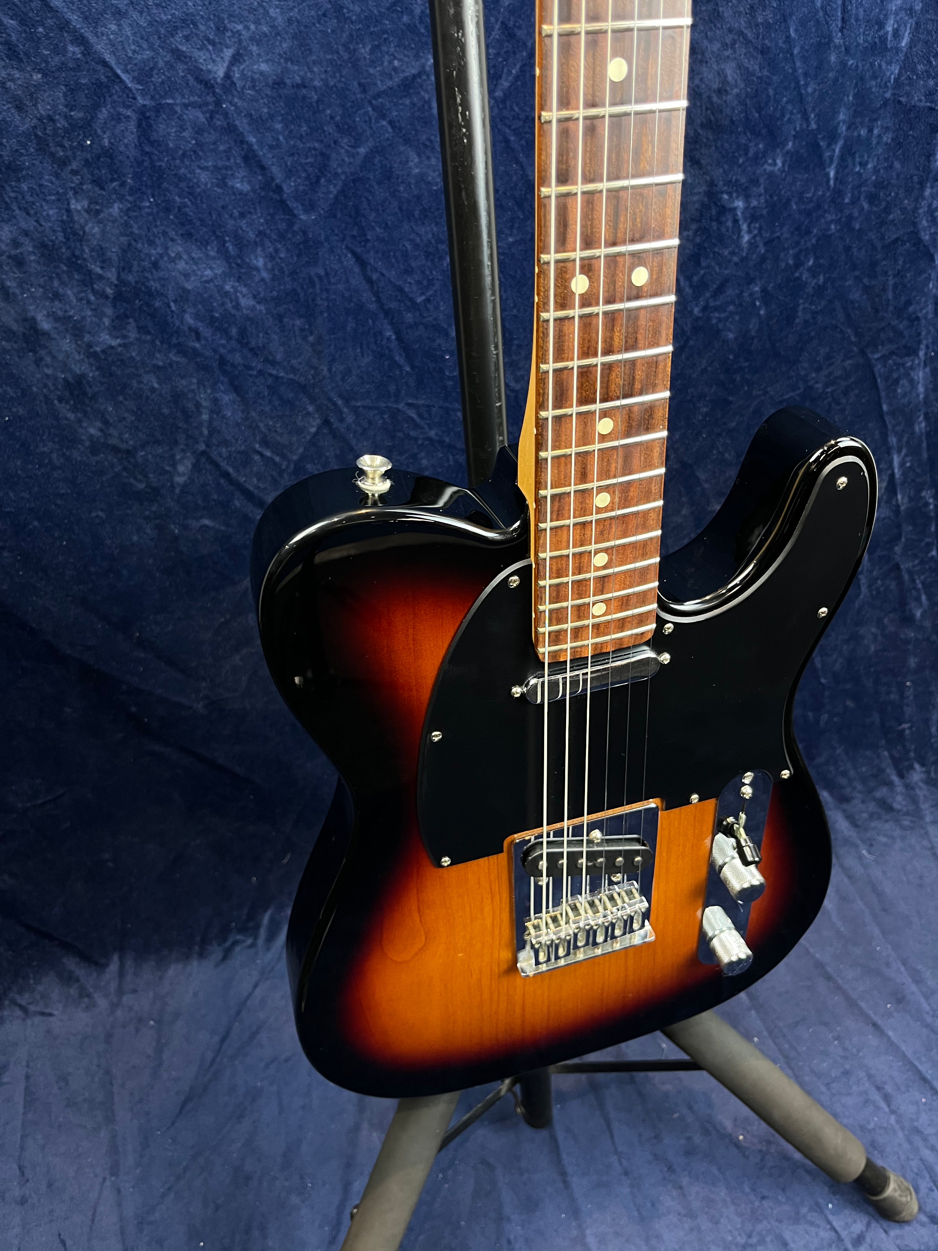 Guitare électrique sunburst avec plaque noire, deux micros, boutons argentés et touche en bois foncé, posée sur un support.