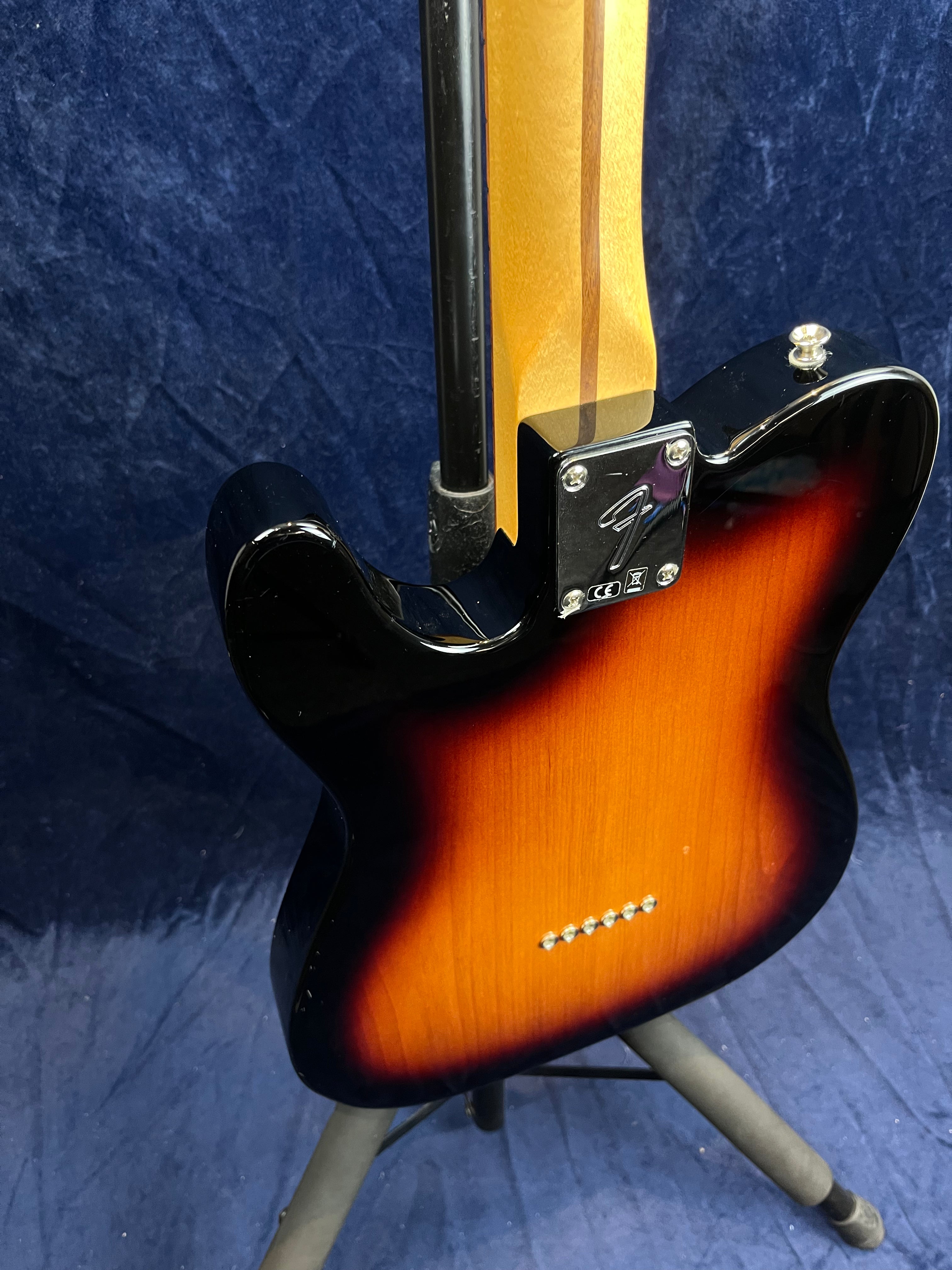 Guitare électrique sunburst vue de dos, manche érable vissé, six points pour cordes traversantes, posée sur un support noir devant un fond bleu.