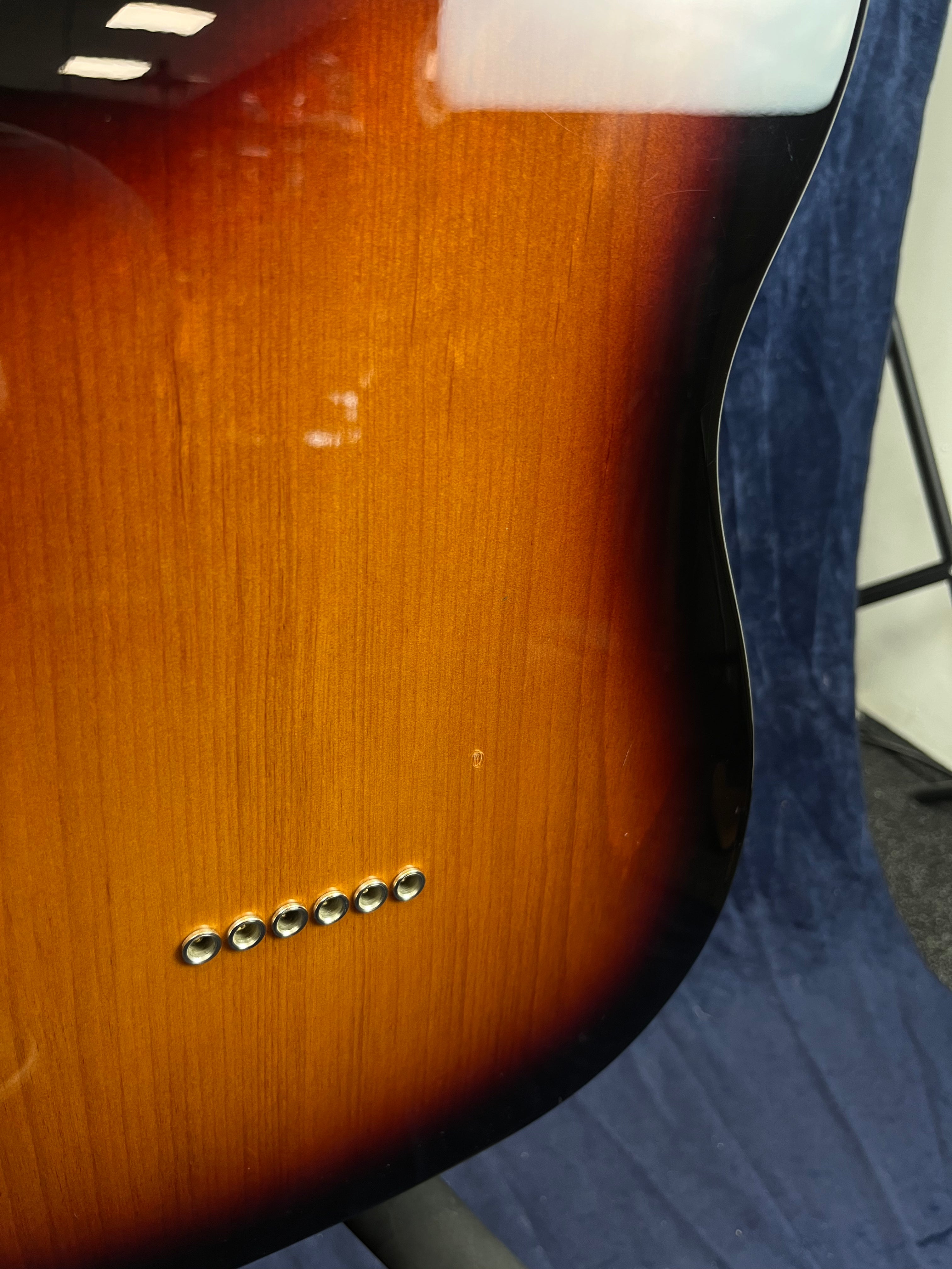 Vernis brillant sunburst sur corps de guitare électrique, six œillets pour cordes visibles en bas à gauche, fond bleu foncé.