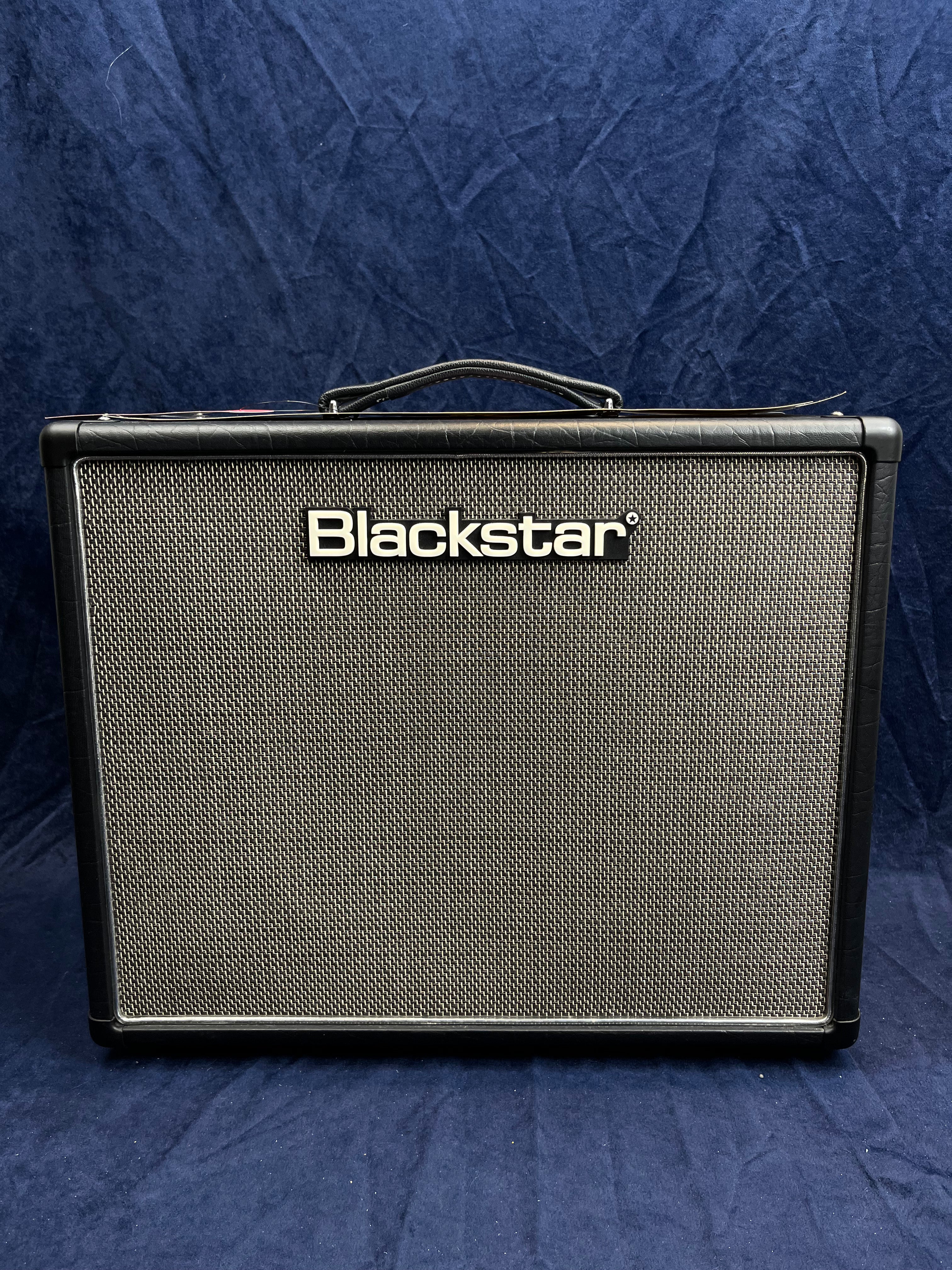 Blackstar amplifier with a black frame, silver mesh front grille, and a carrying handle on top, placed against a dark blue background.