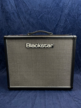 Blackstar amplifier with a black frame, silver mesh front grille, and a carrying handle on top, placed against a dark blue background.