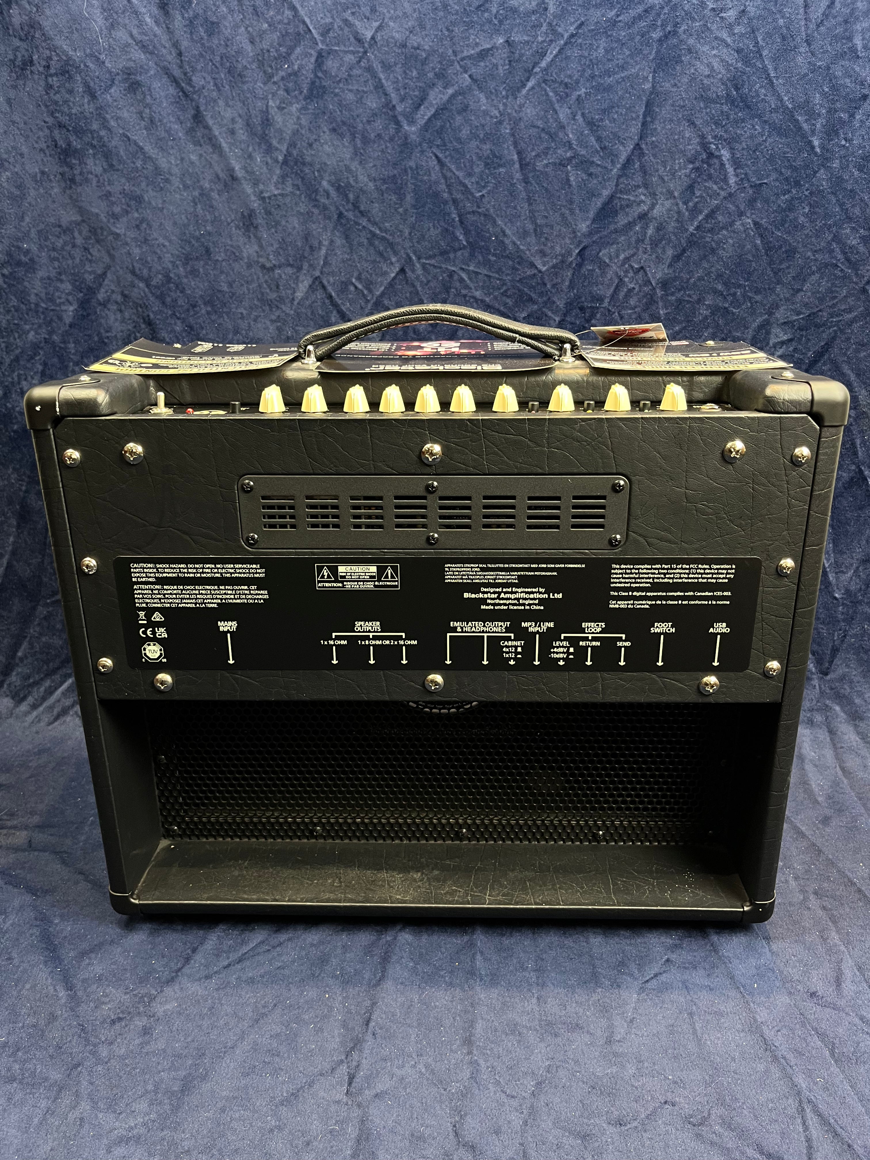 Black guitar amplifier viewed from the back, showing a row of cream control knobs on top, a carry handle, multiple input/output jacks, and warning labels.