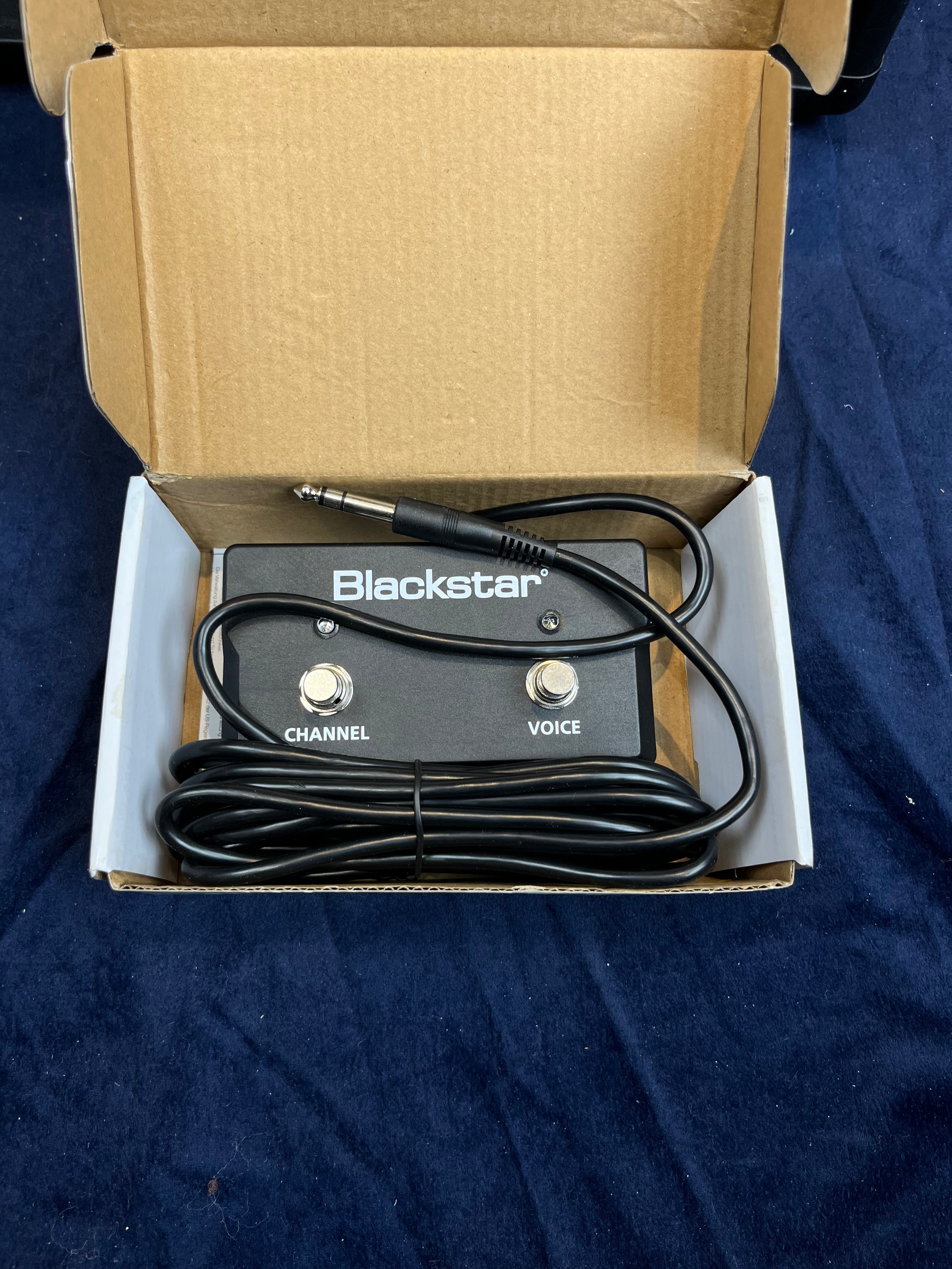 Blackstar amplifier footswitch with CHANNEL and VOICE buttons, connected cable, and a 1/4-inch jack plug, placed in an open cardboard box.