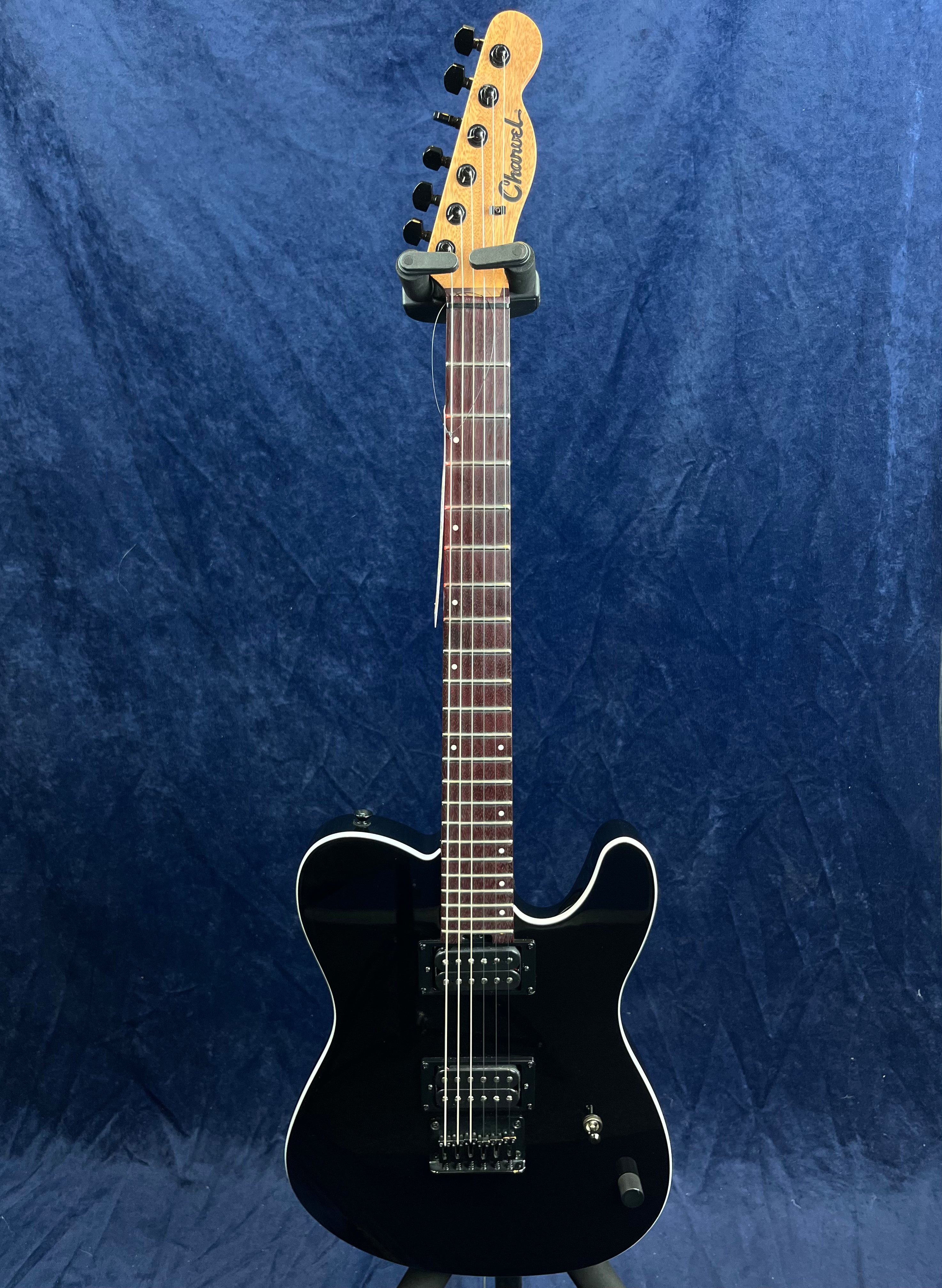 Black Charvel electric guitar with white trim, two humbucker pickups, rosewood fretboard, and single cutaway body, standing upright against a blue background.