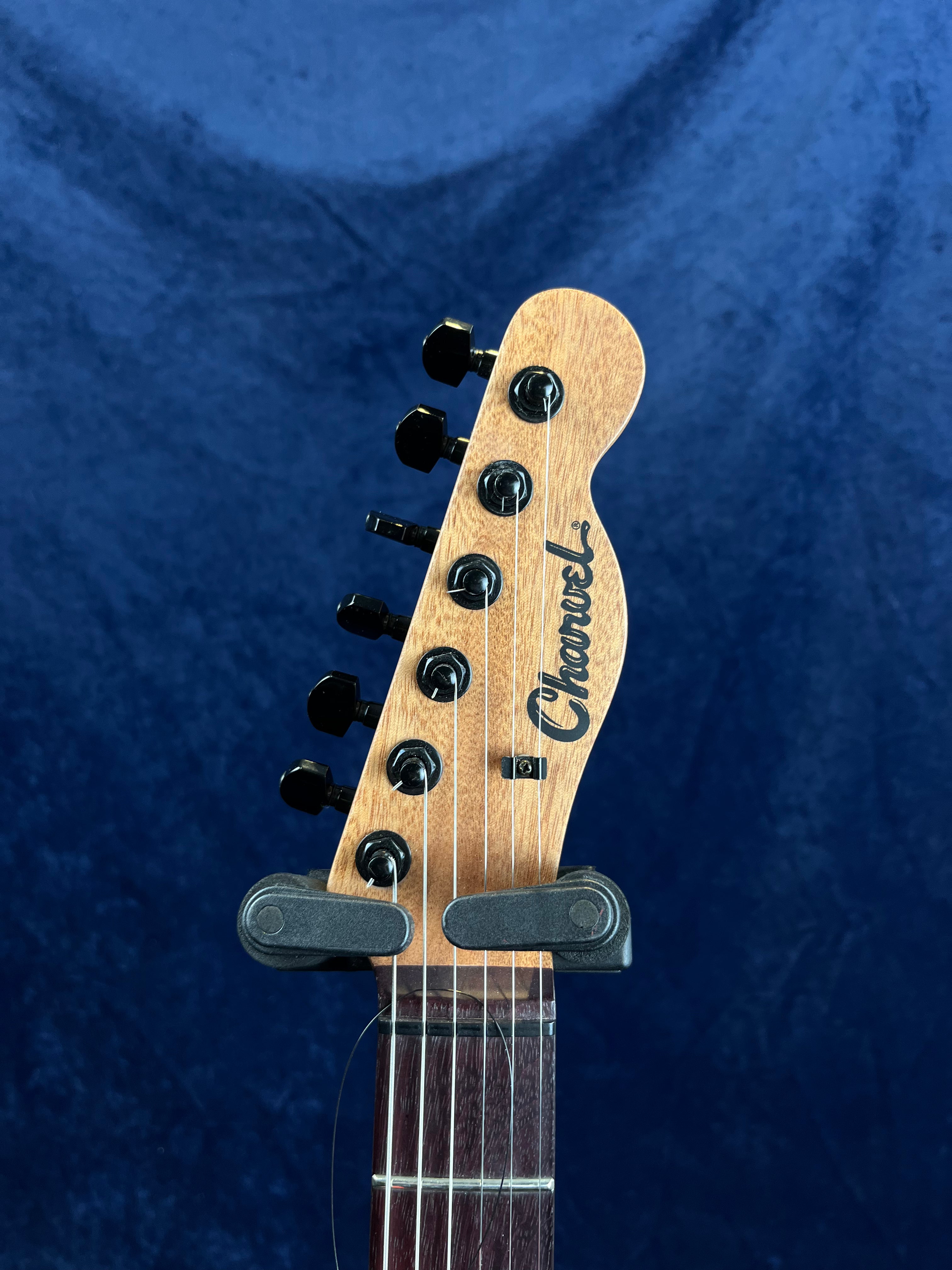 The headstock of a Charvel electric guitar with six black tuning pegs and a string retainer bar, set against a dark blue background.