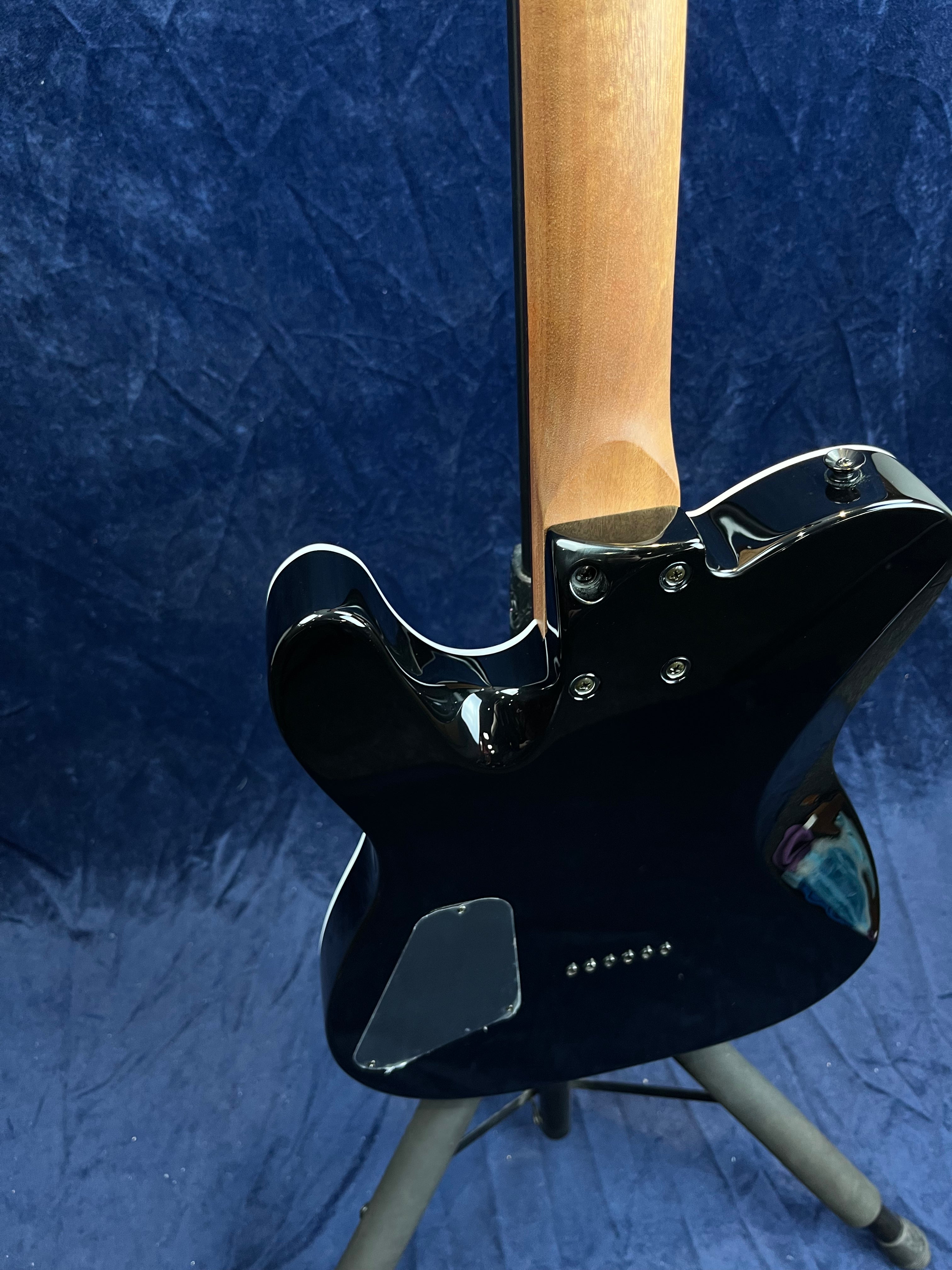 Black electric guitar shown from the back, with a natural wood neck and a rectangular rear electronics cover plate, resting on a stand against a dark blue background.