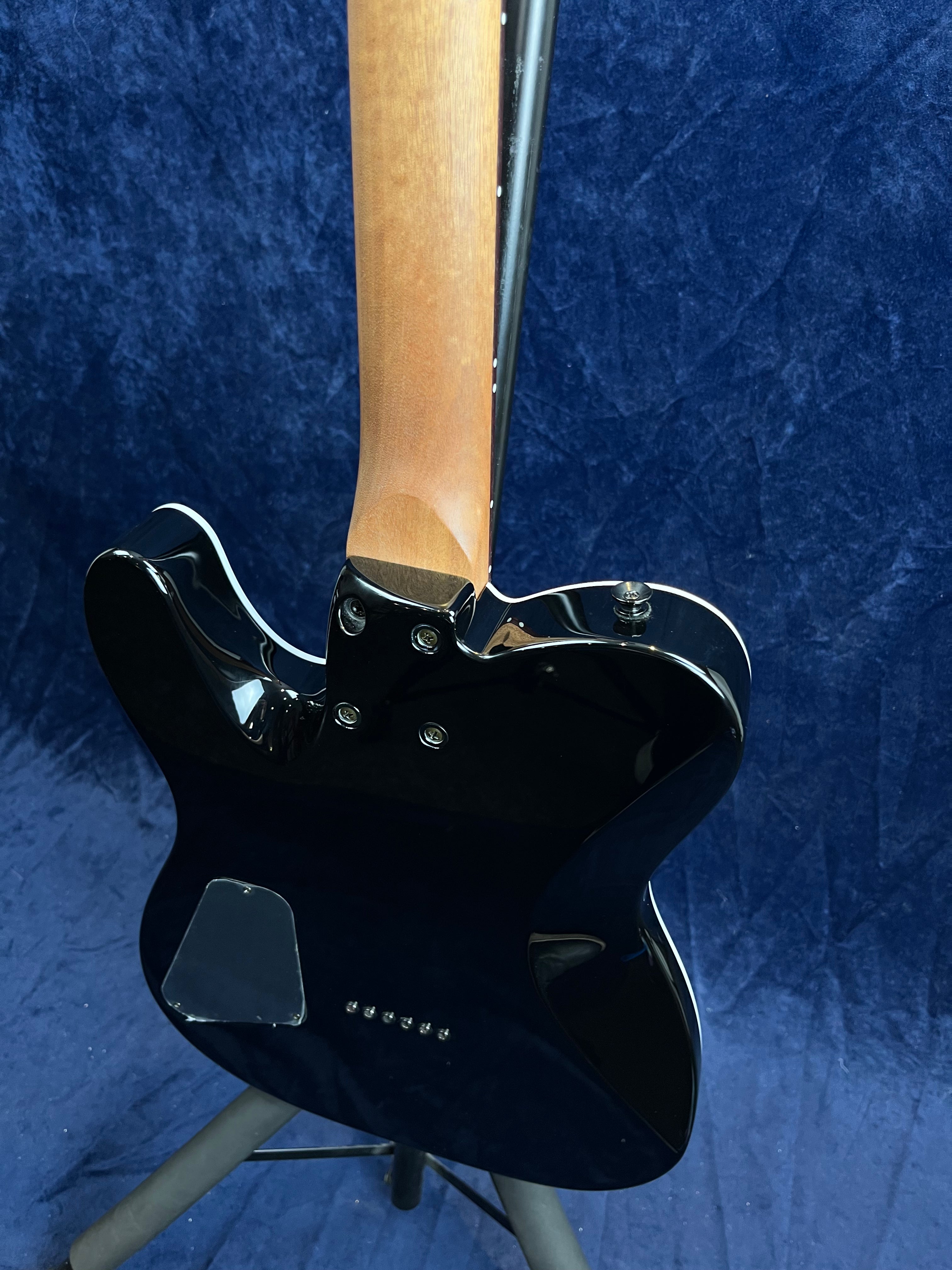 Black electric guitar seen from the back, with a natural wood neck and metal bolts attaching the neck to the body, standing on a tripod stand.