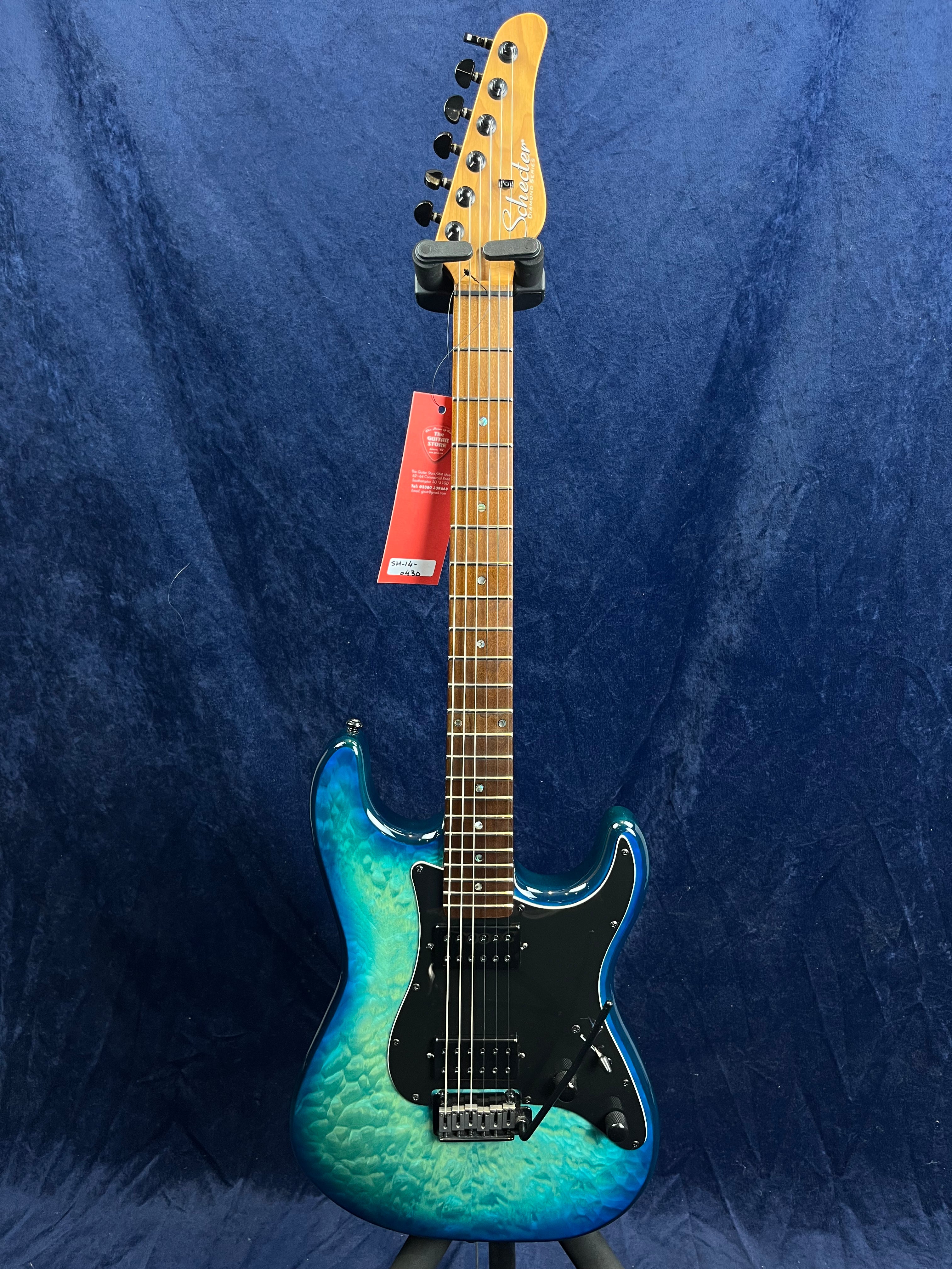 Schecter electric guitar with a blue-green quilted finish, black pickguard, maple neck, six tuning pegs, and a red tag hanging from the headstock.