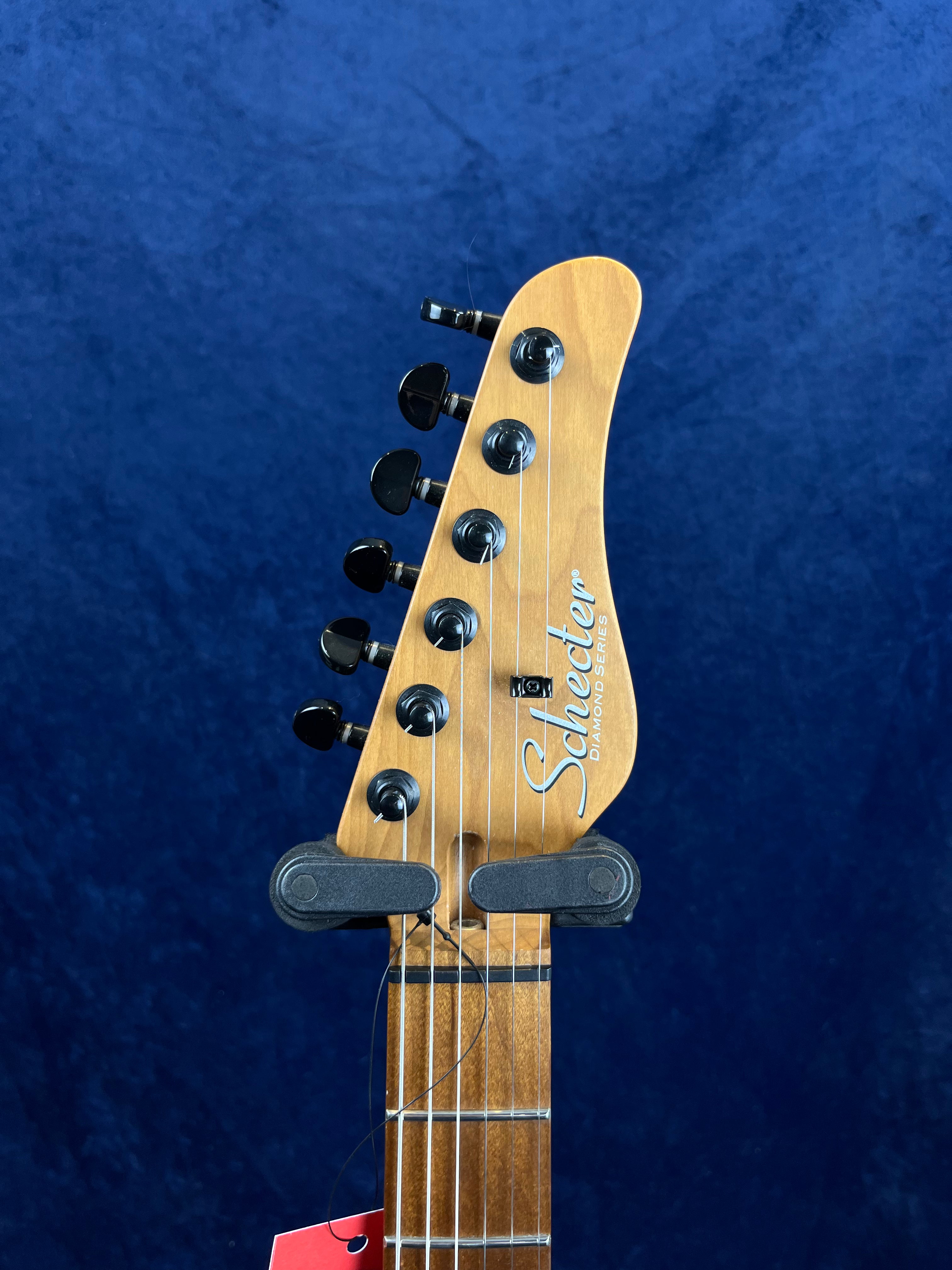 Schecter Diamond Series electric guitar headstock with six black tuning pegs and strings, set against a blue background.