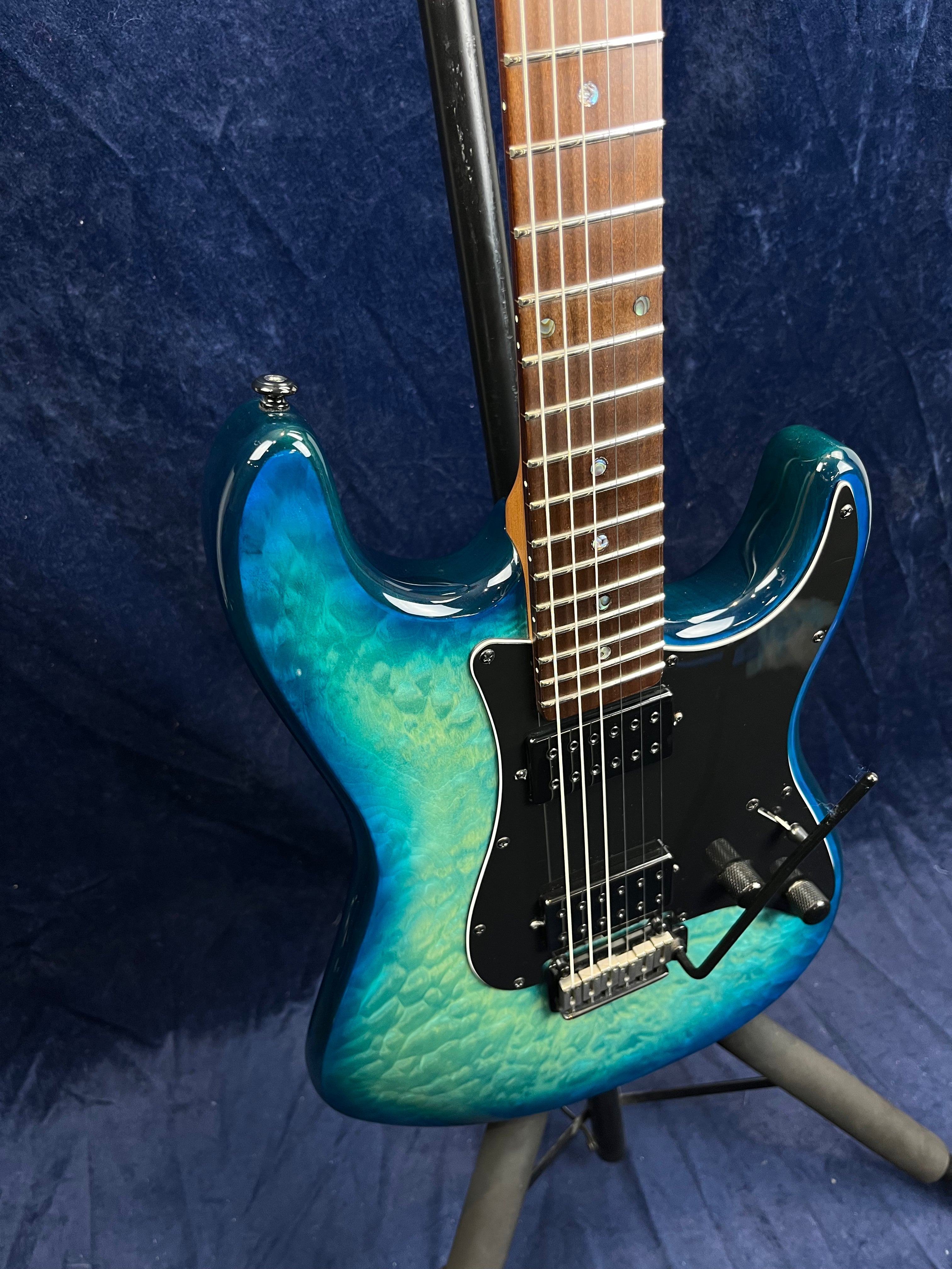 Blue-green electric guitar with quilted maple top, two humbucker pickups, black pickguard, whammy bar, and rosewood fretboard on a stand.