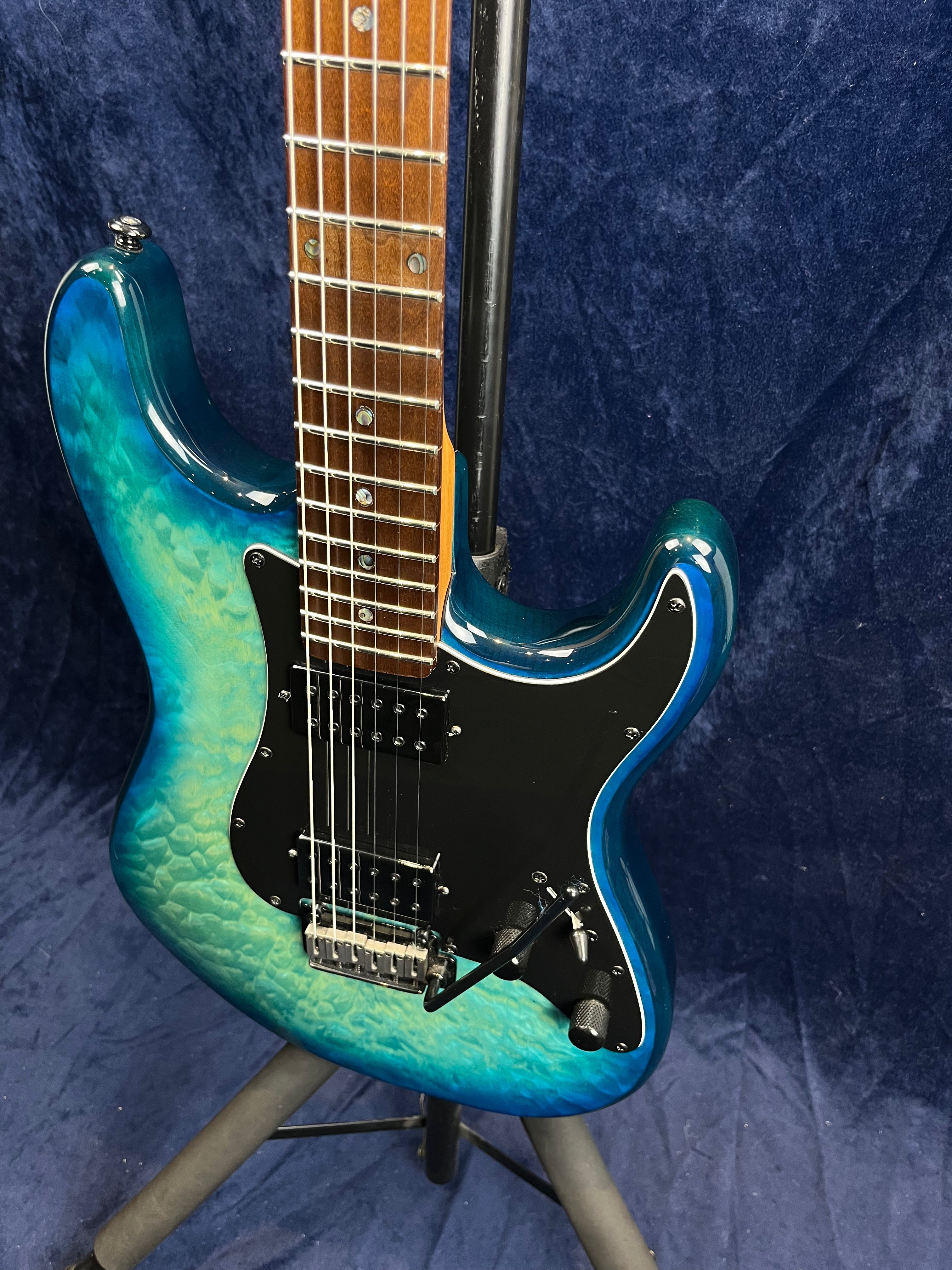 Electric guitar with a blue-to-green quilted finish, black pickguard, two humbucker pickups, and a tremolo bridge, shown on a stand.