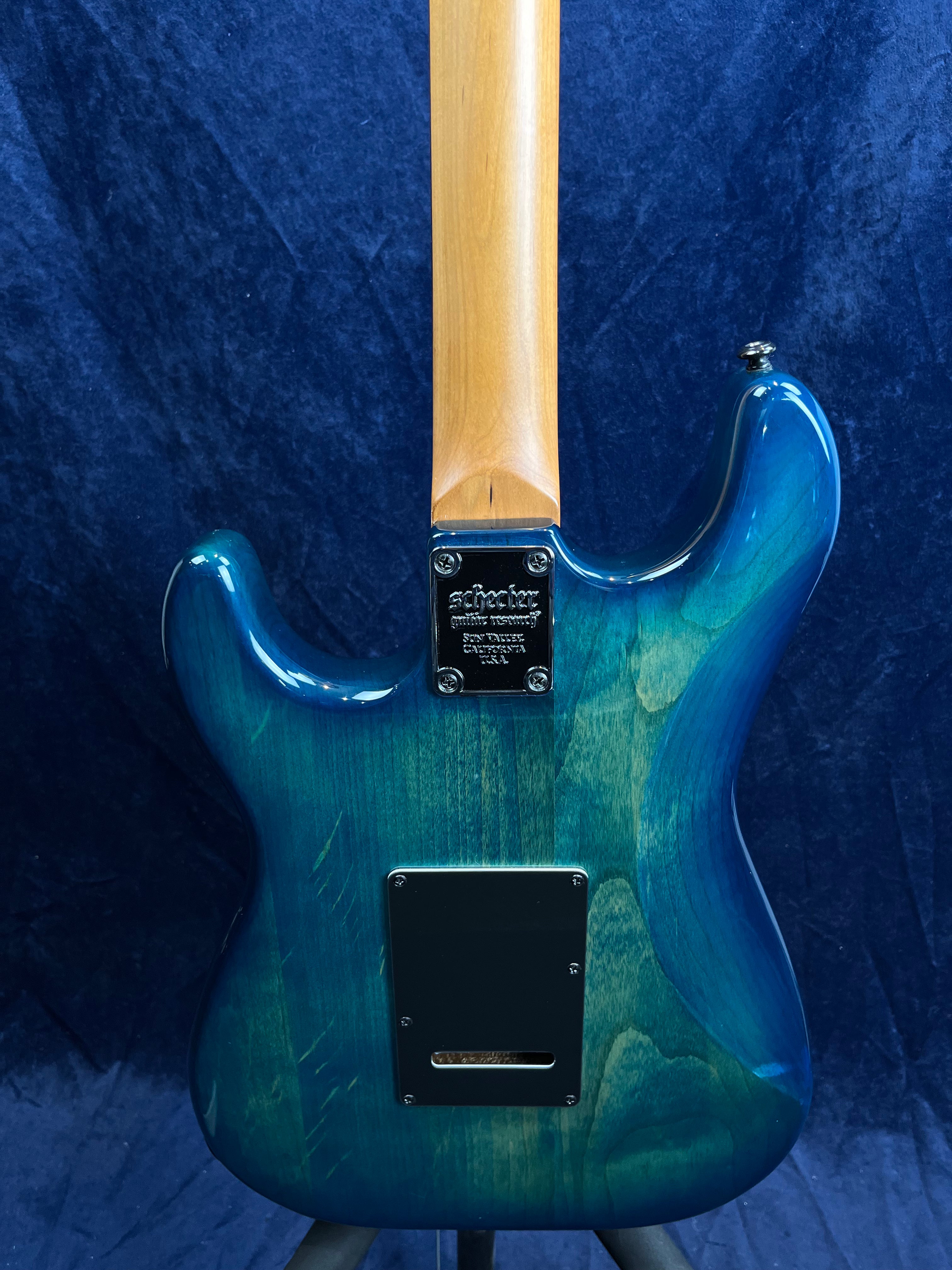 Blue-green Schecter electric guitar viewed from the back, showing the wooden neck, metal neck plate, and a rectangular electronics cover plate.