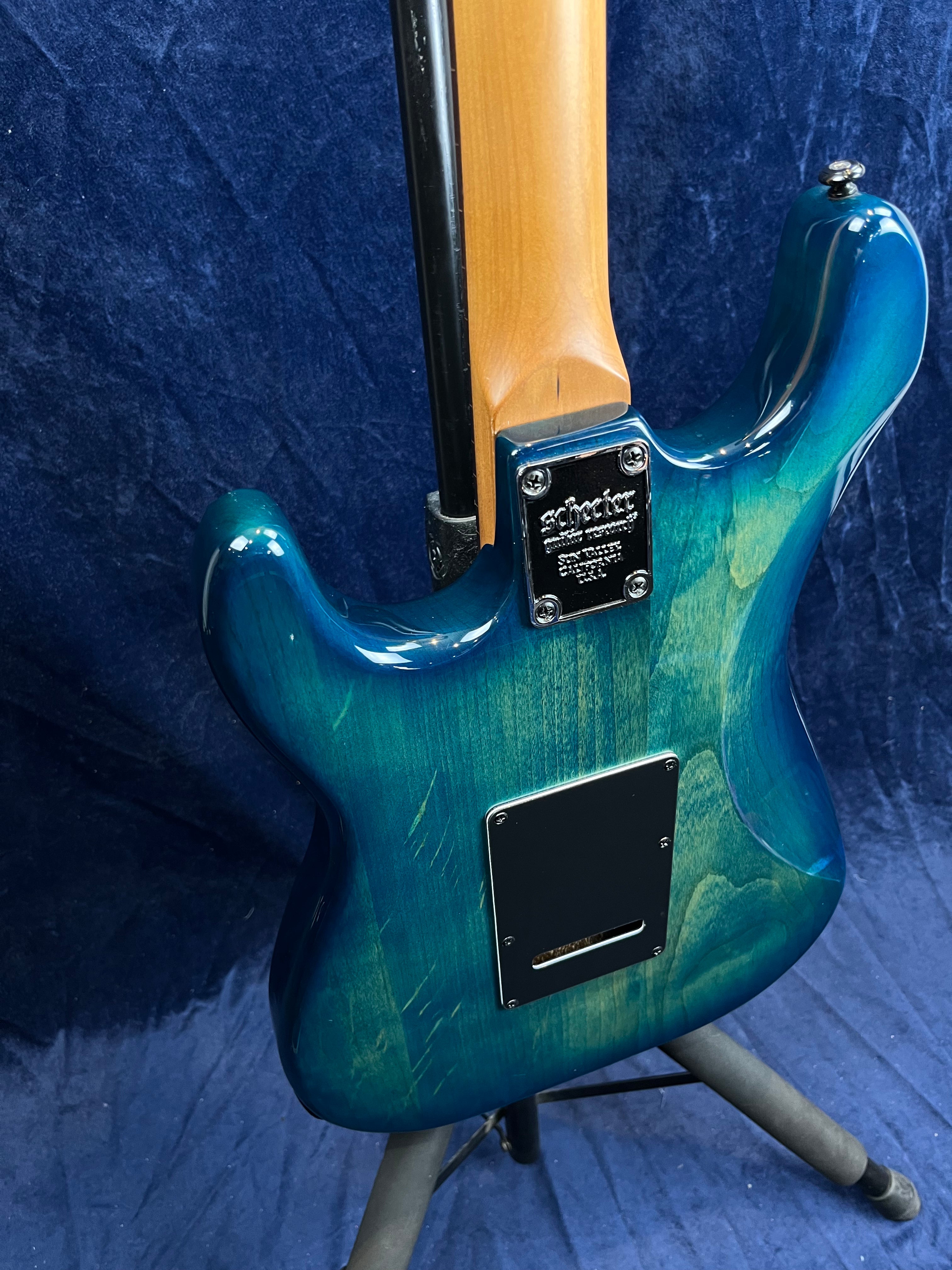 A Schecter electric guitar with a glossy blue-green finish shown from the back, revealing the neck joint and rear cavity cover plate.
