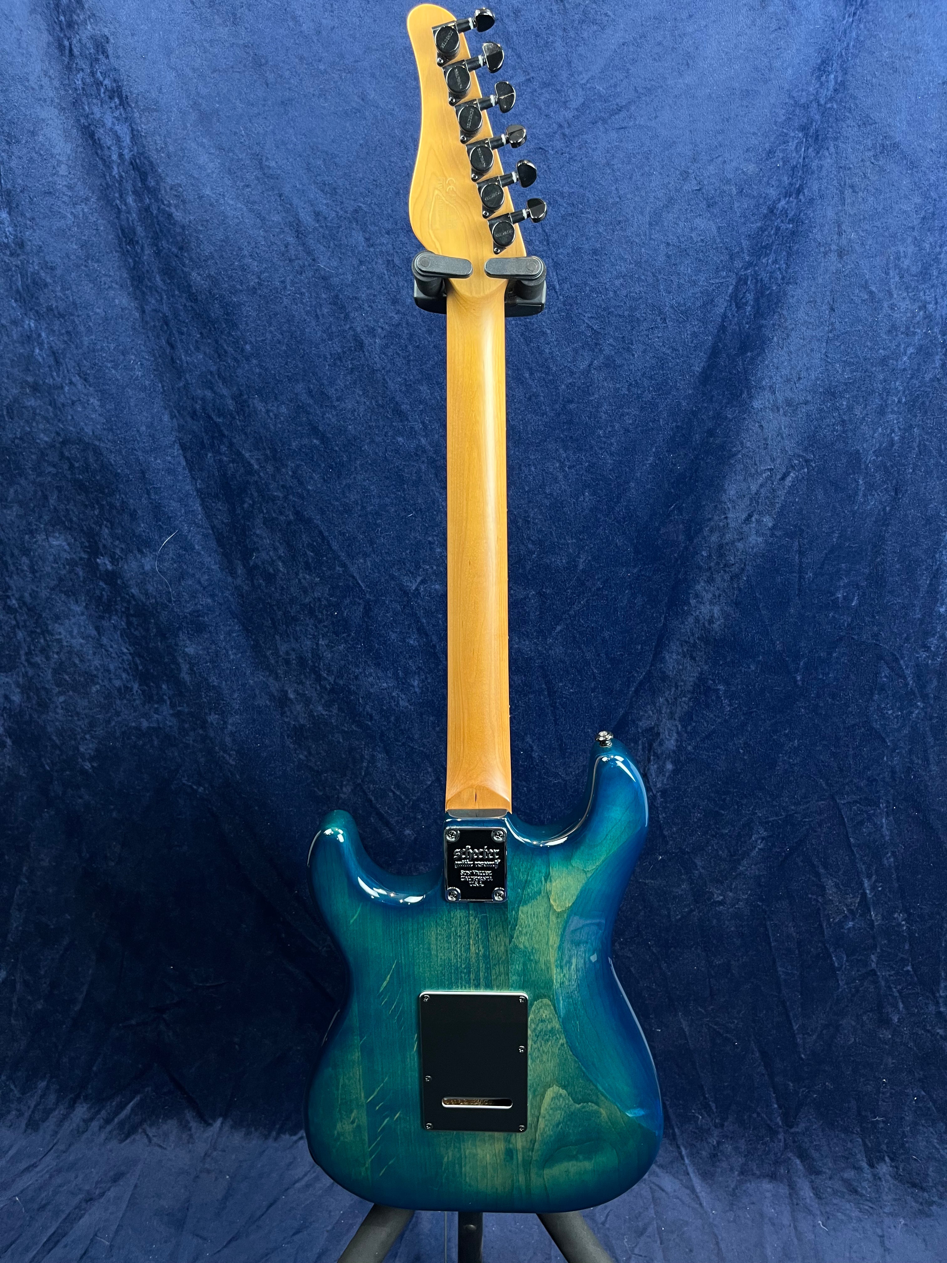 A blue-green electric guitar seen from the back, showing a natural wood neck and black tuning pegs against a blue fabric background.