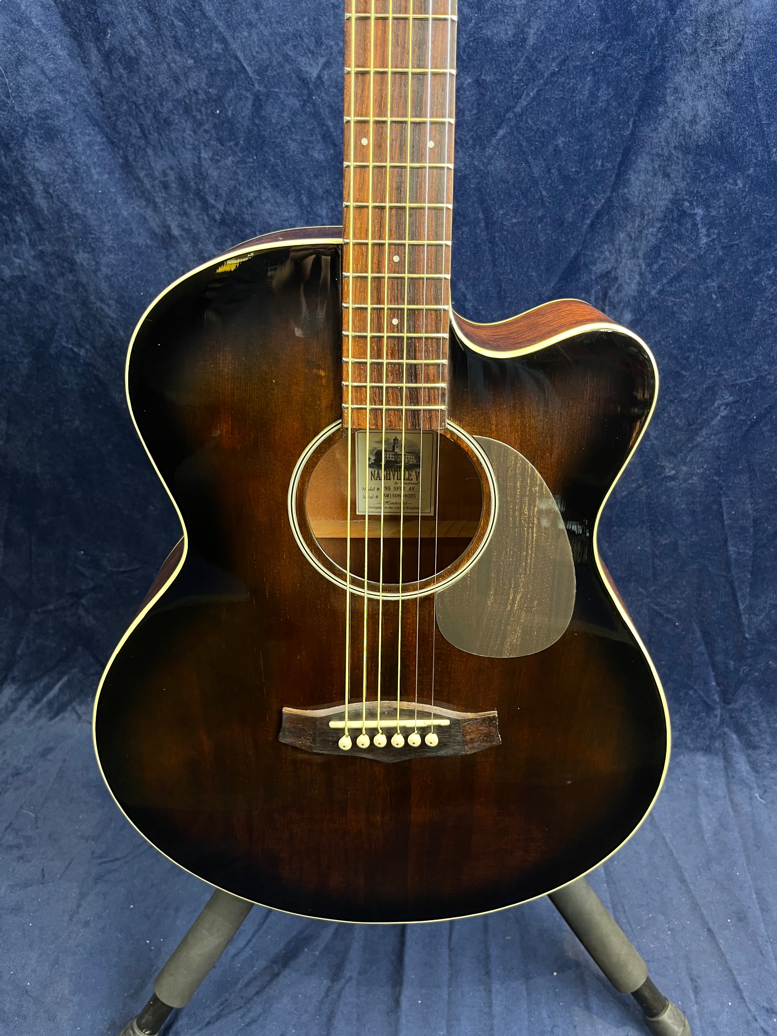 Dark brown acoustic guitar with a cutaway body, six strings, cream edge binding, and a dark pickguard, shown upright on a stand against a blue background.
