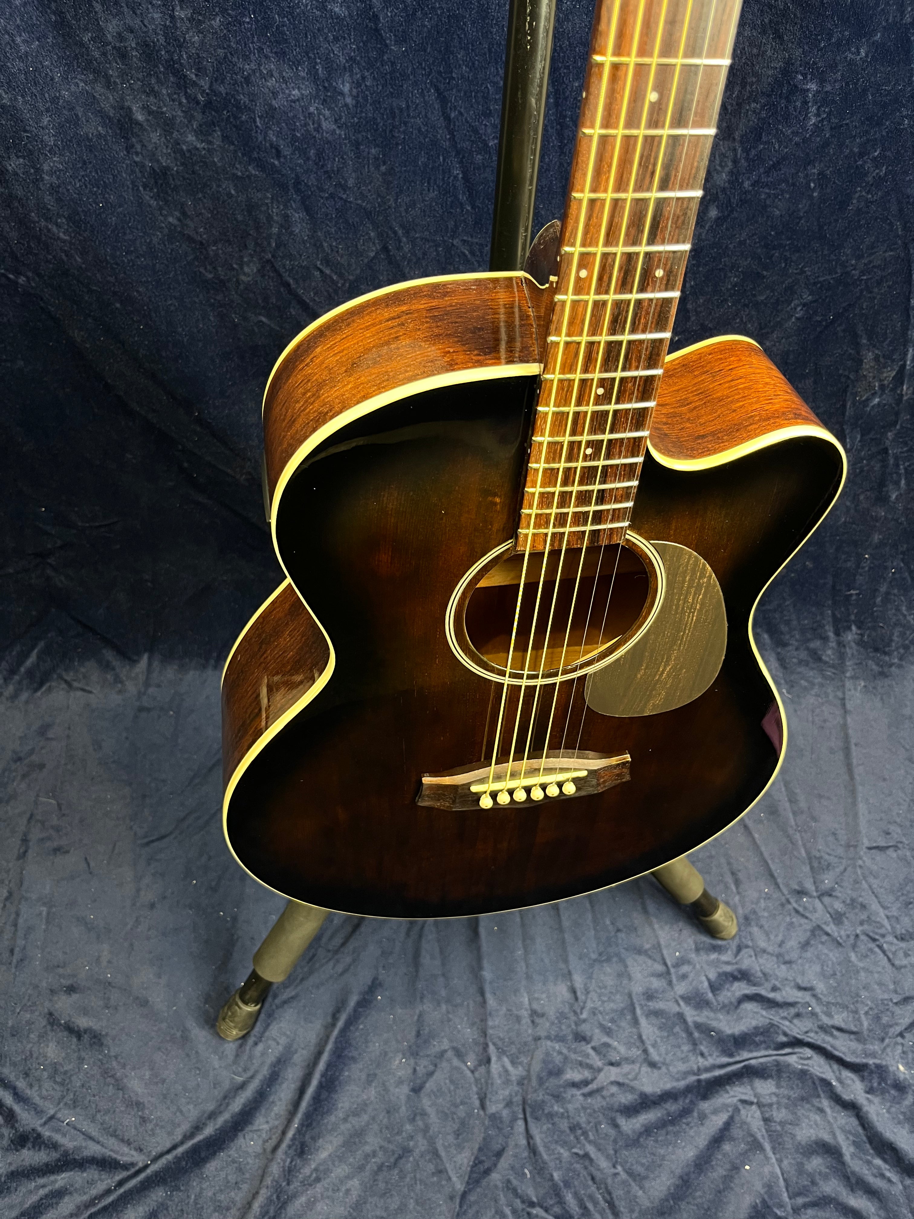 Dark brown acoustic guitar with light trim, six strings, and a rounded cutaway body, resting on a stand against a dark fabric background.