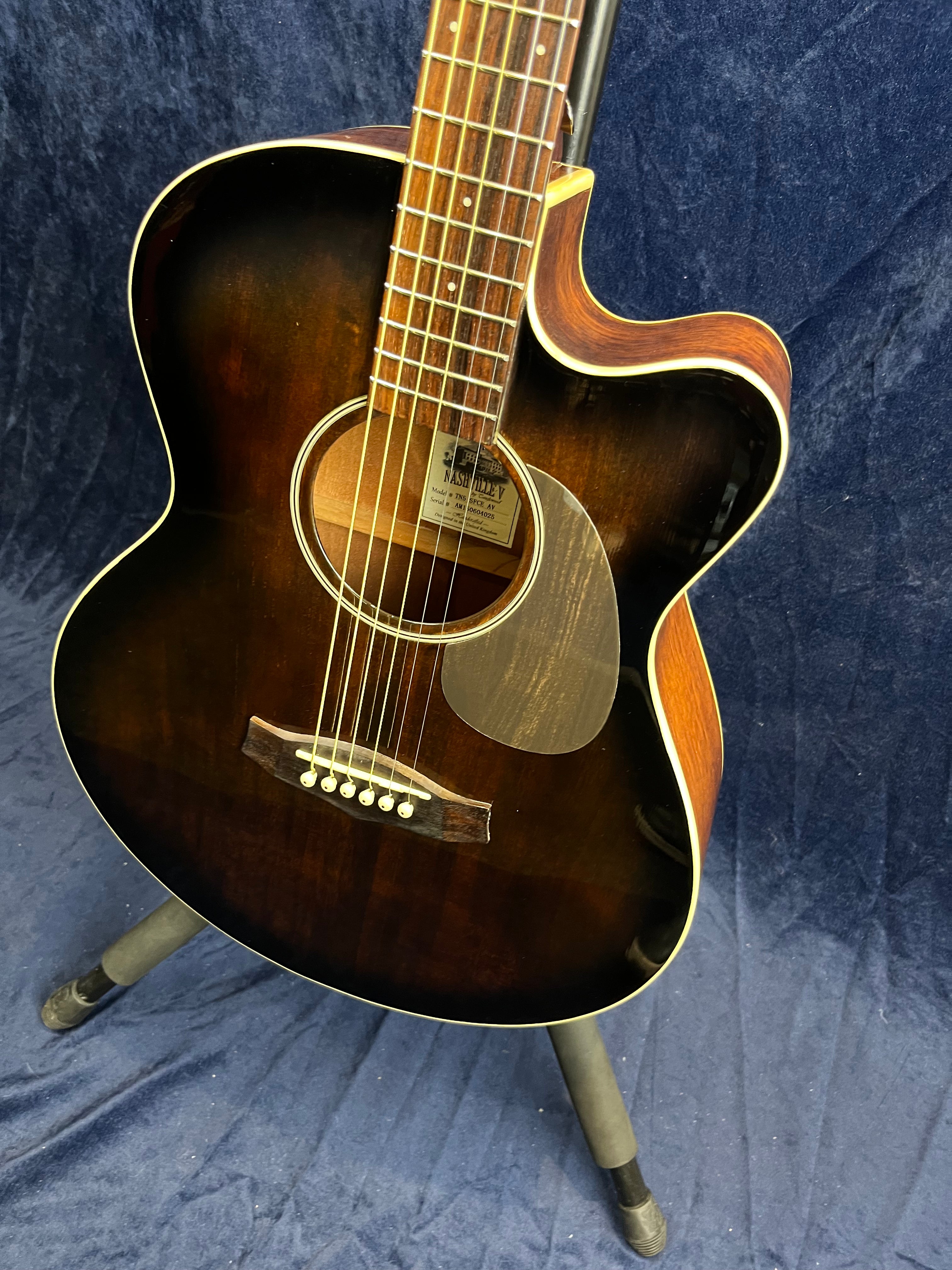 Dark sunburst acoustic guitar with a cutaway body, six strings, wooden bridge, and a tortoiseshell pickguard, standing on a tripod stand.