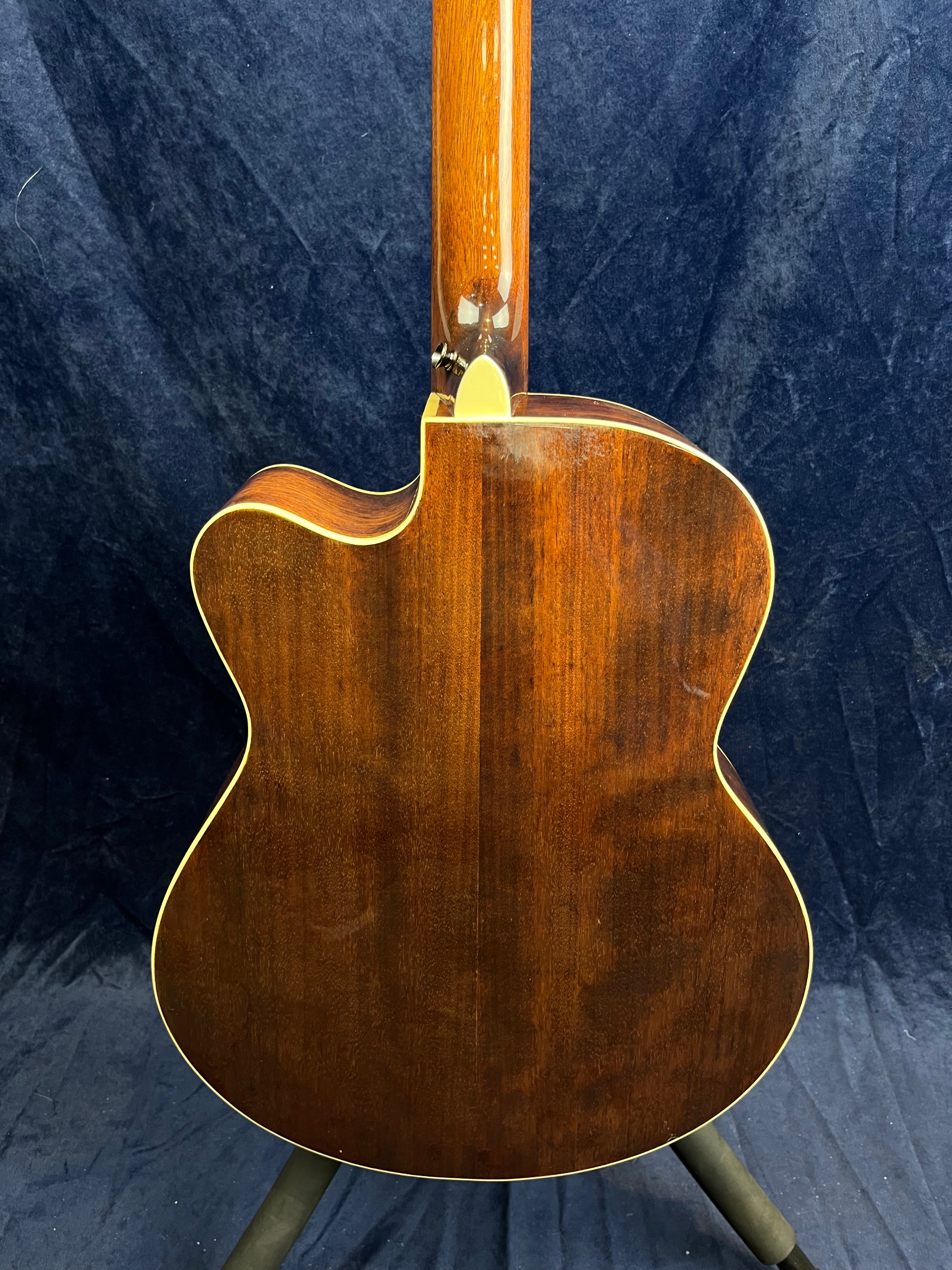 Back view of an acoustic guitar with dark wood finish, cream binding, and a cutaway body shape, displayed on a stand against a dark background.