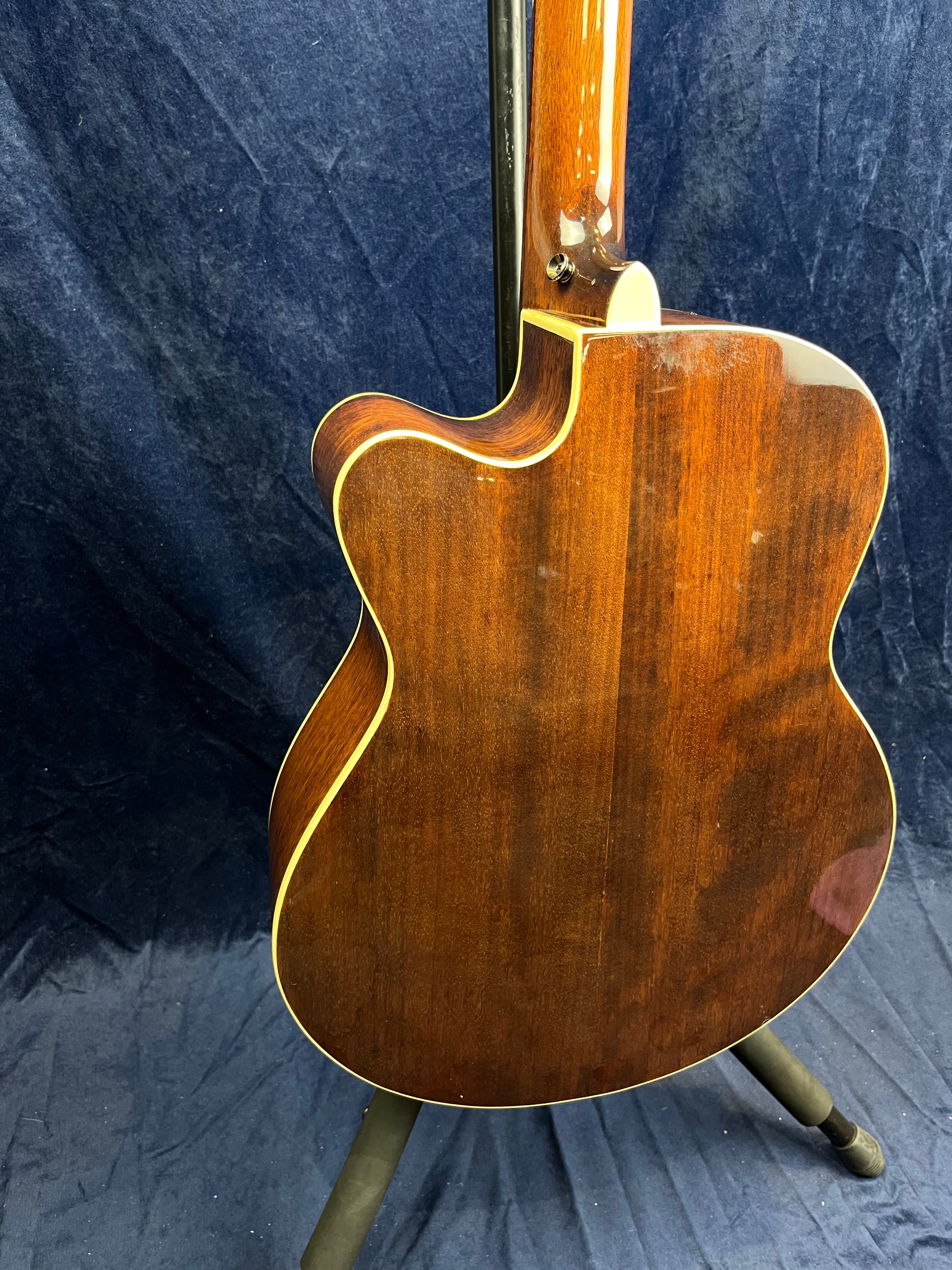 Glossy brown acoustic guitar back with cream binding, curved cutaway, and slight scuff marks near the neck joint, resting on a stand.