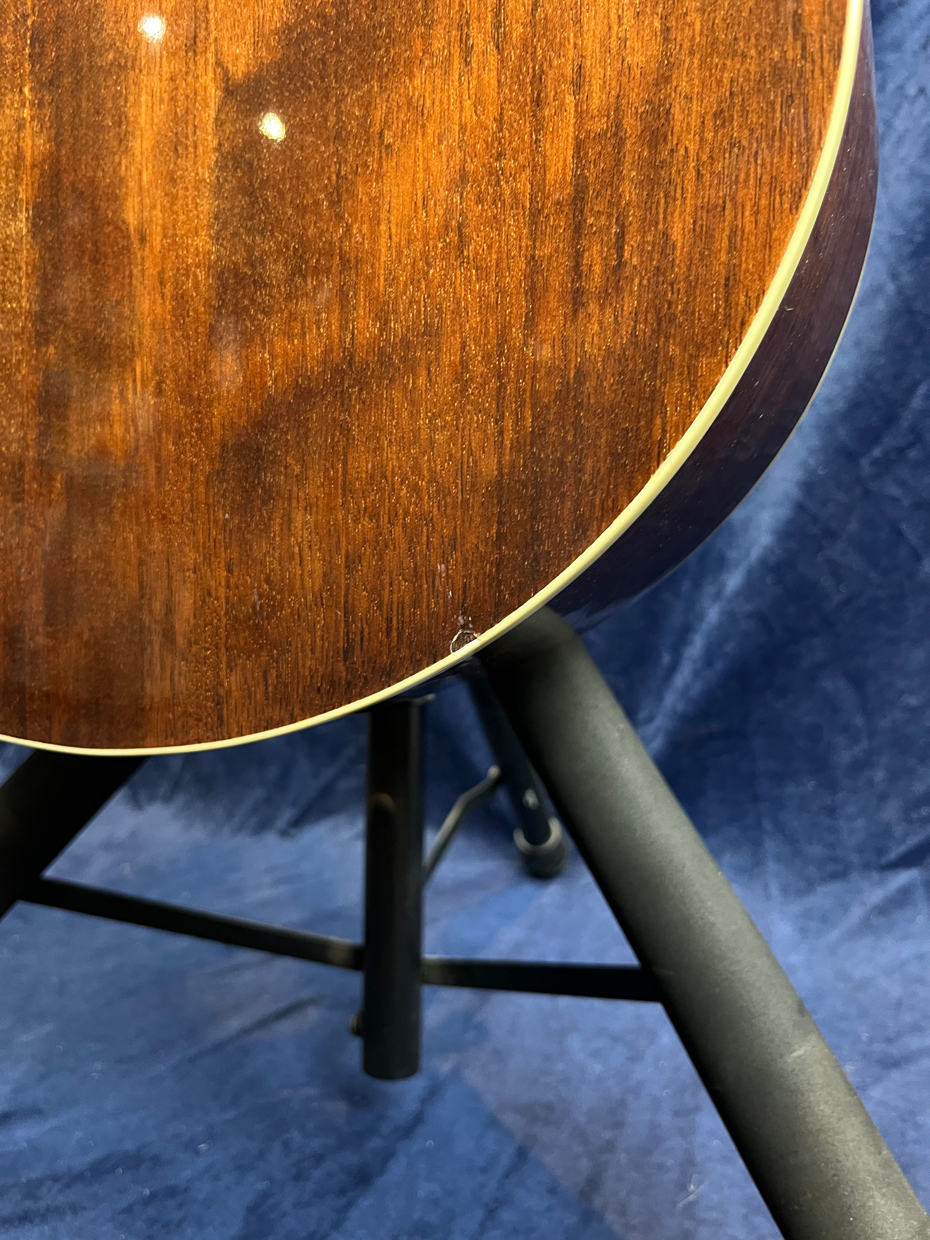 Lower side of a brown acoustic guitar body showing a small chip on the edge, with cream binding and part of a black guitar stand visible.