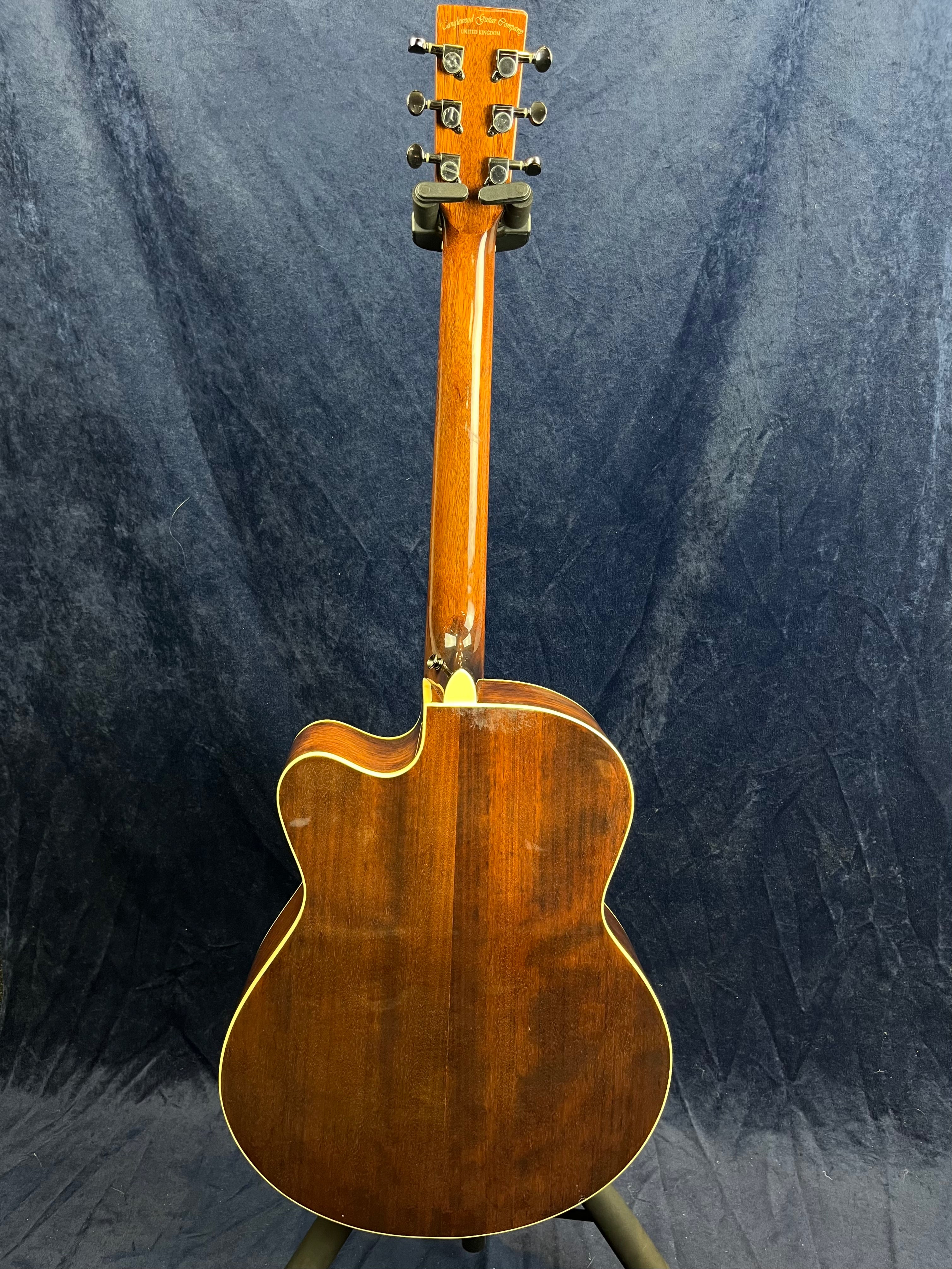 Back view of an acoustic guitar with a glossy dark wood finish, cream binding, and six tuning pegs, displayed on a stand against a dark fabric background.