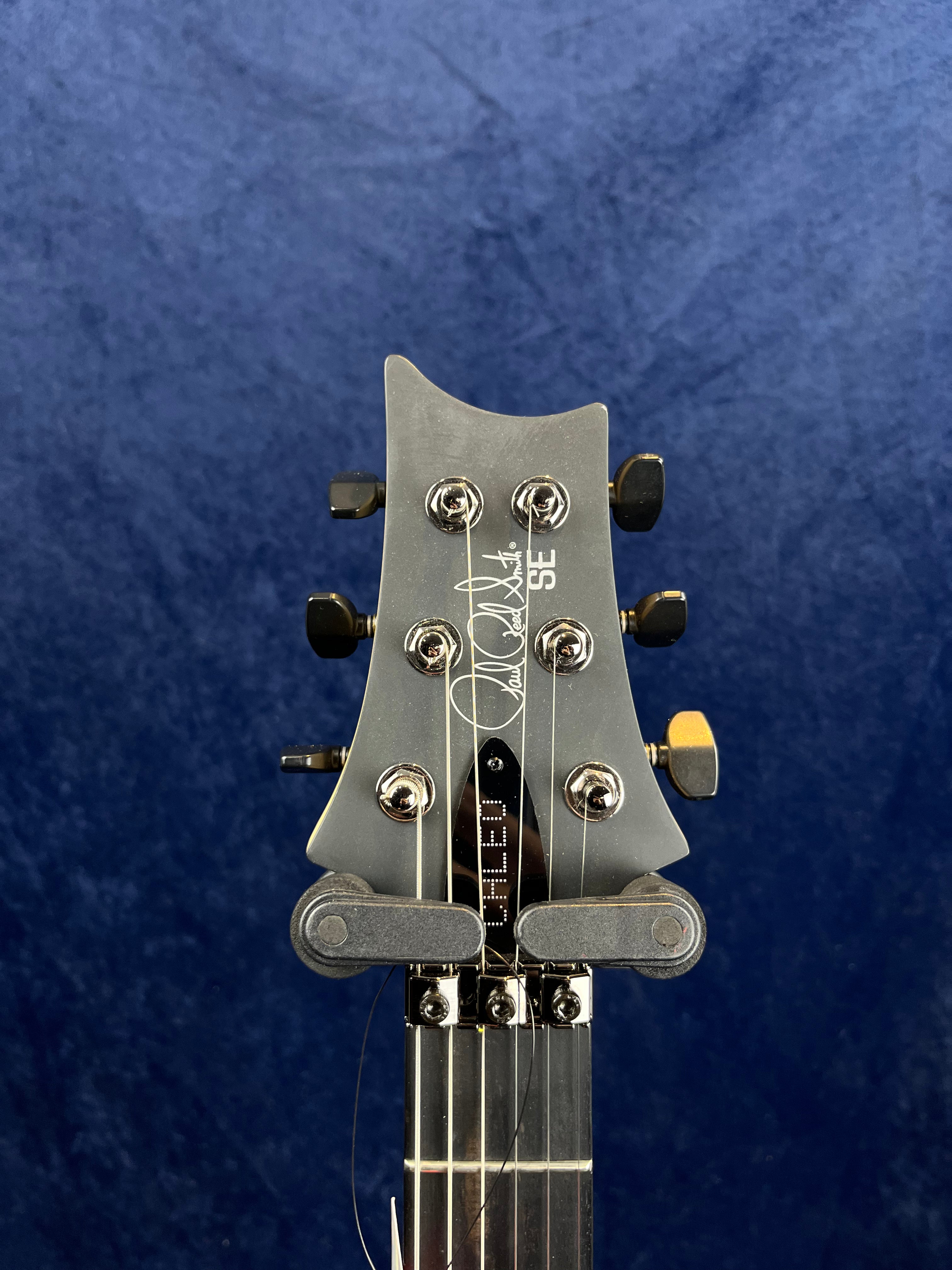Headstock of an electric guitar with six tuning pegs, signature, "SE" logo, and a capo clipped below the tuners, set against a blue background.