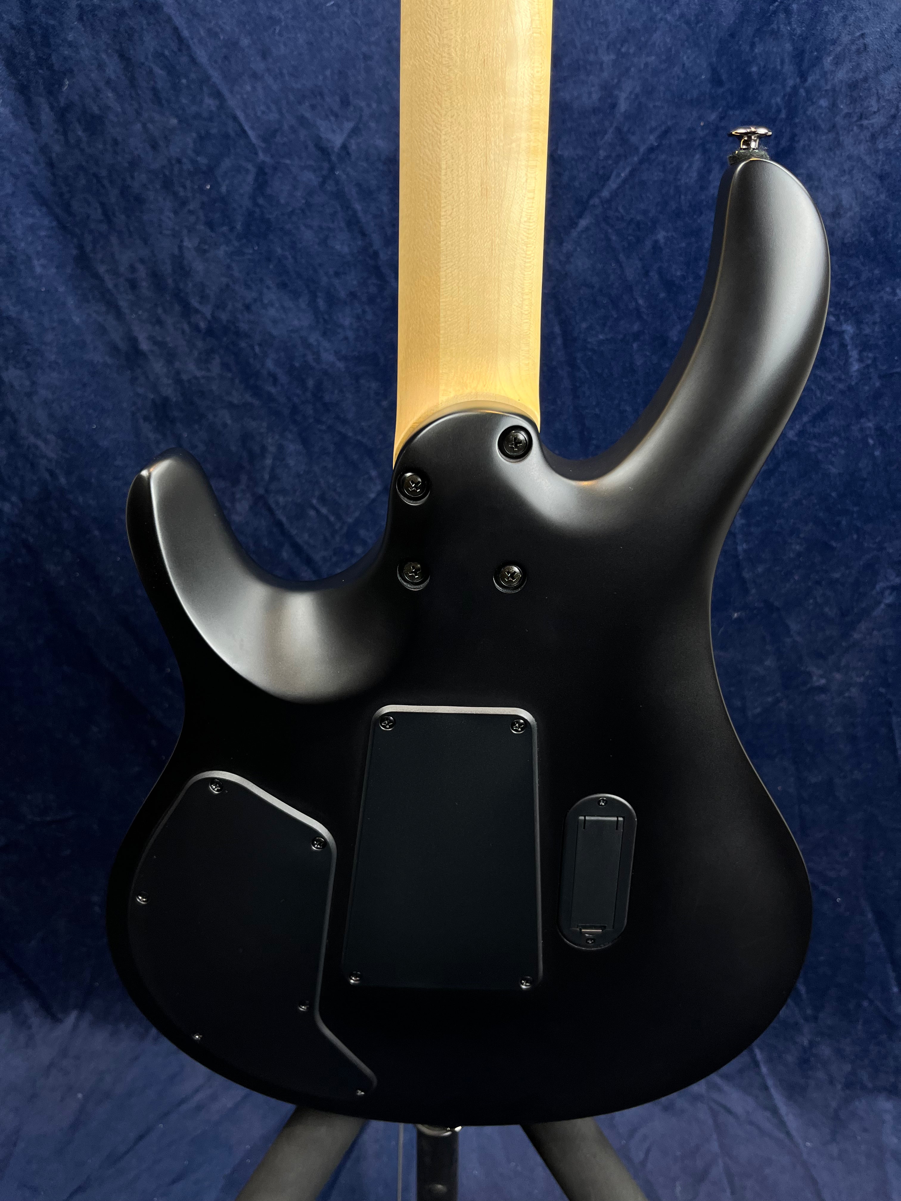 Matte black electric guitar back showing three electronics panels and a light-colored neck against a dark blue background.