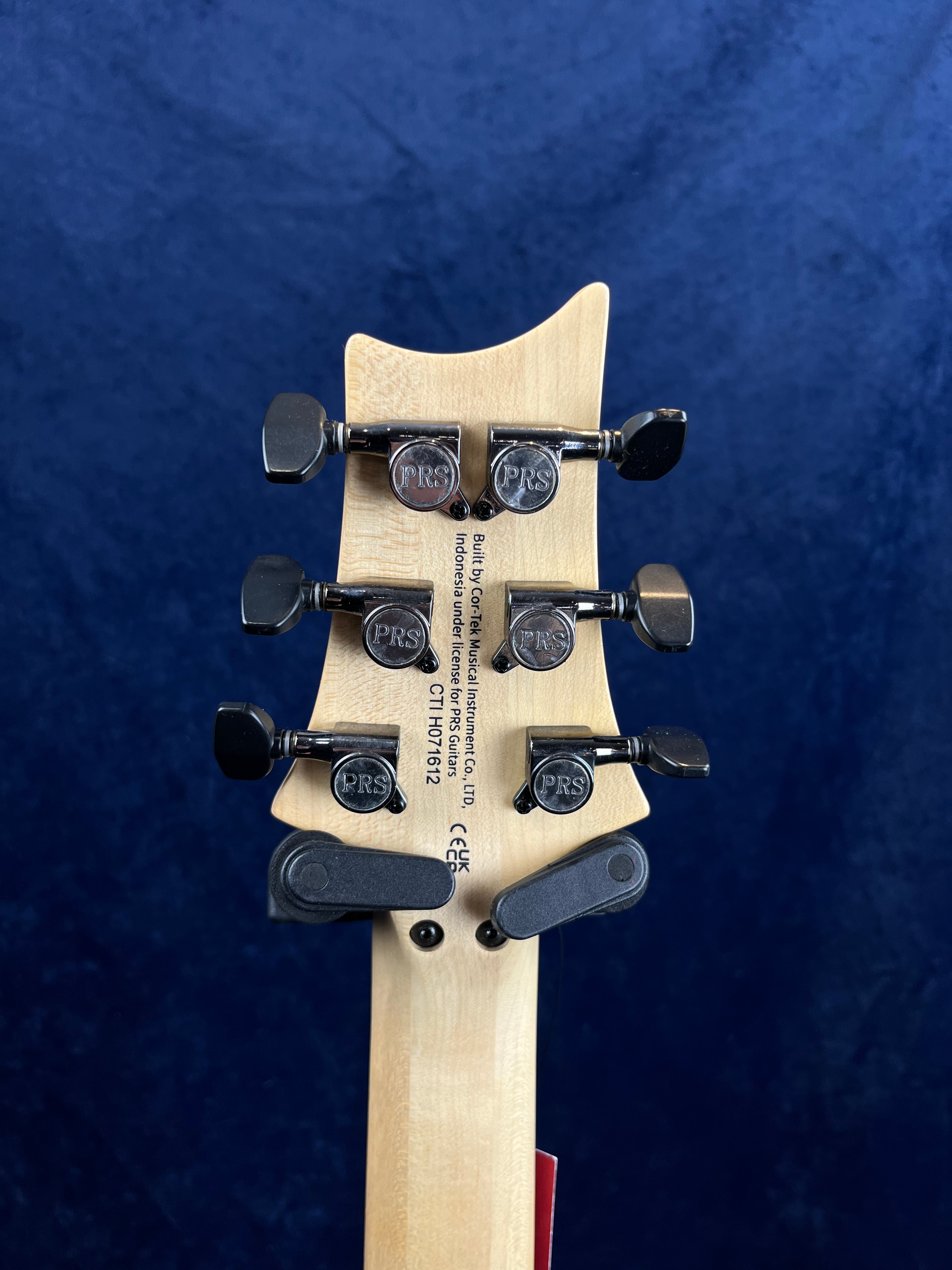 Back of a PRS electric guitar headstock showing six black tuning pegs, PRS logos, serial number, and manufacturer info on light-colored wood.