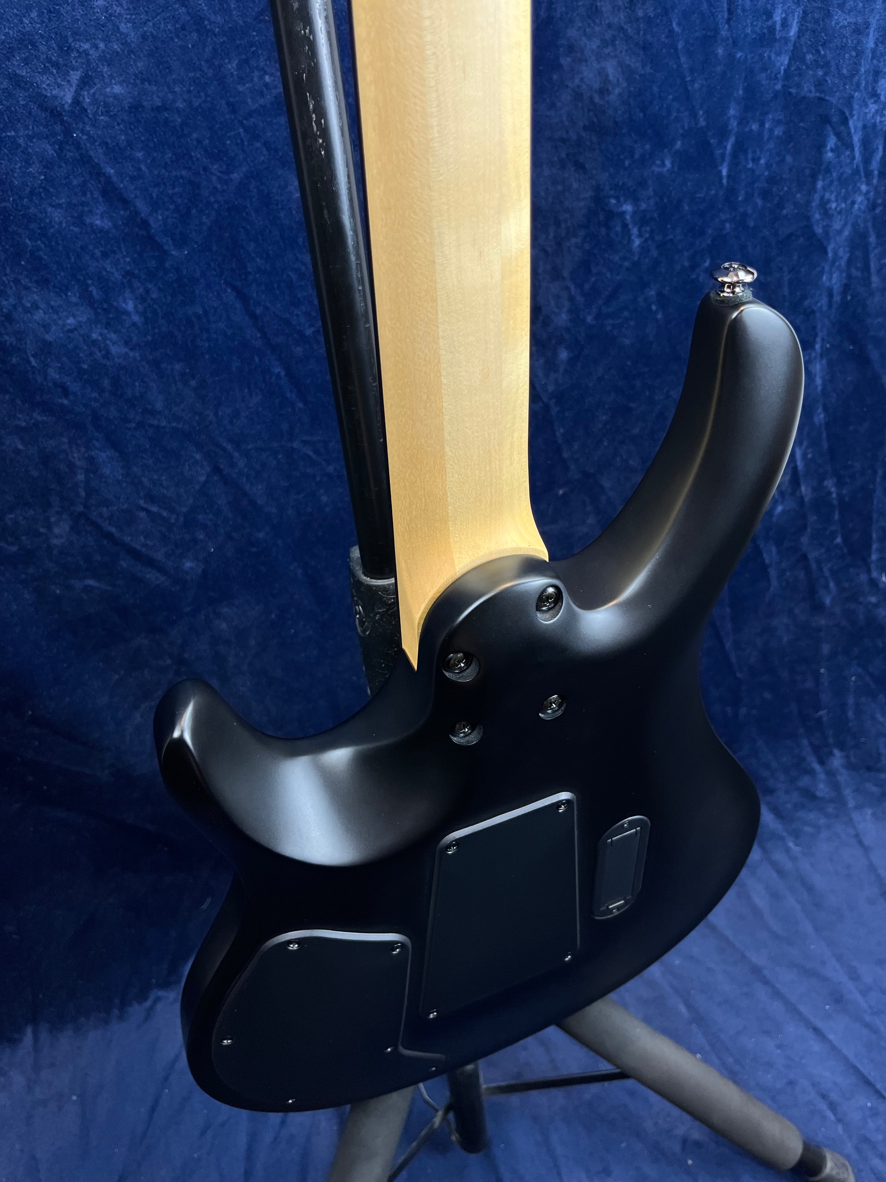 Matte black electric guitar viewed from the back, showing a light wood neck, attached electronics panels, and metallic strap button on a stand.