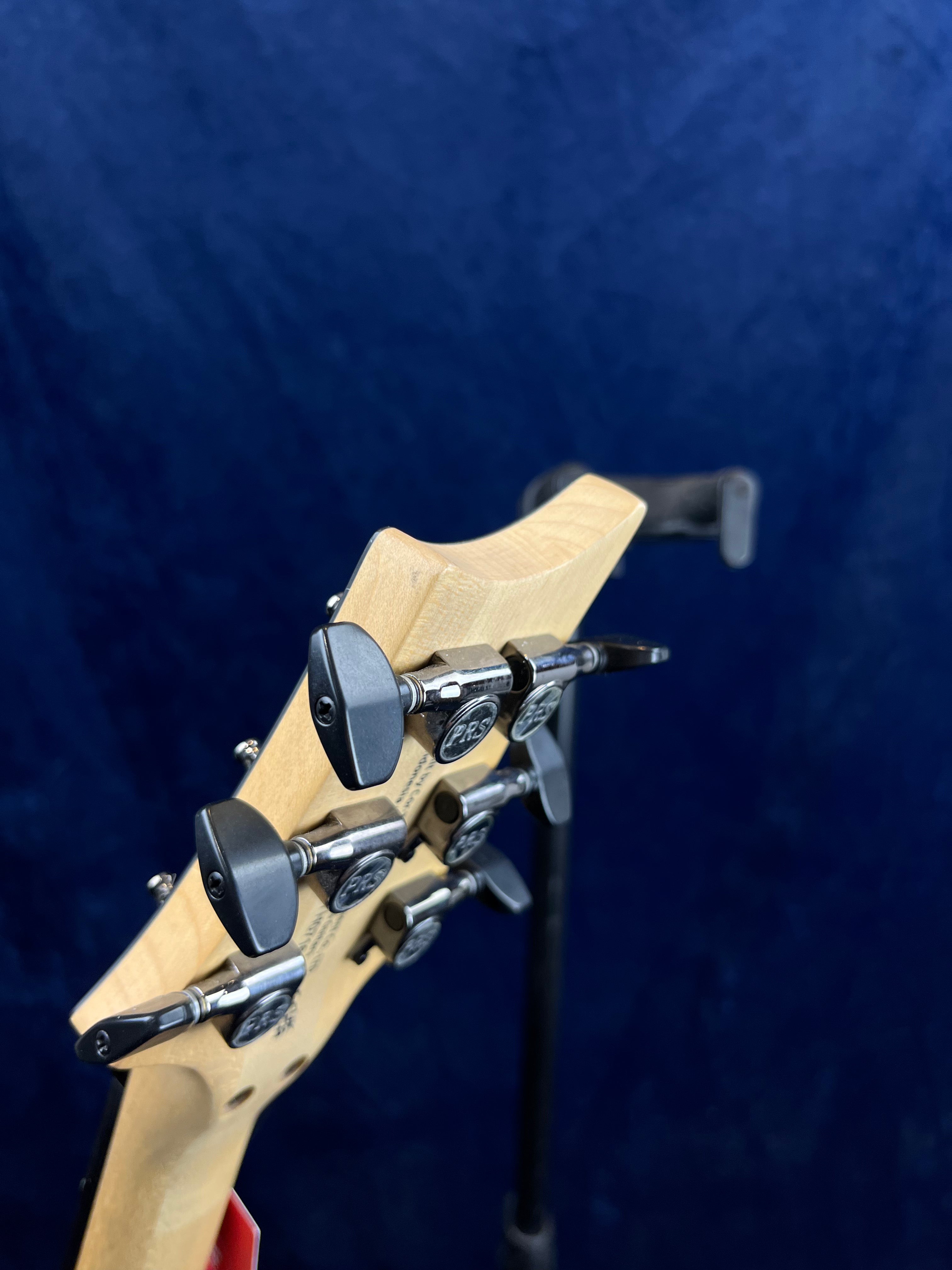 Six tuning pegs marked "PRS" on the back of a natural wood electric guitar headstock, with a black guitar stand against a dark blue background.