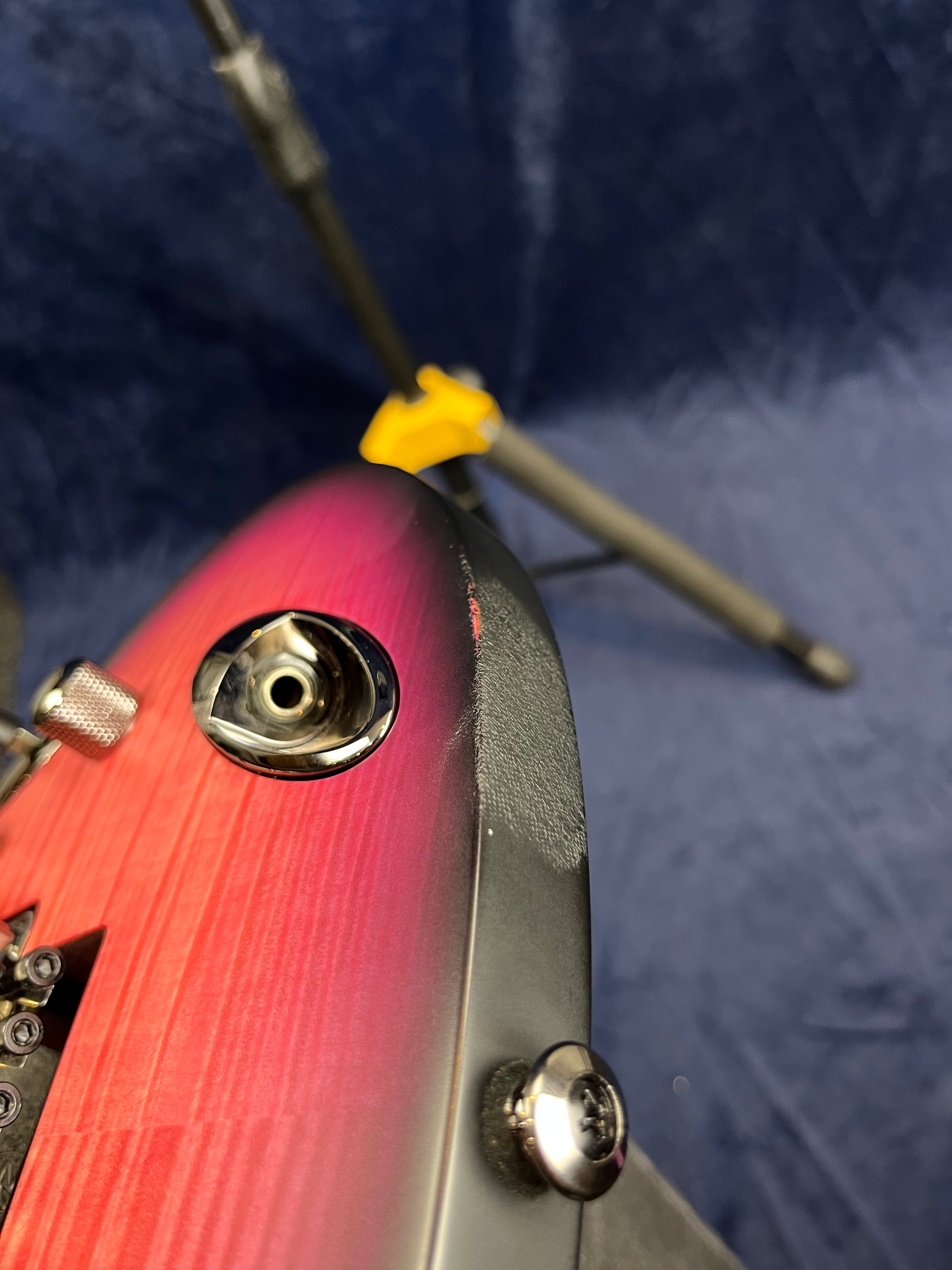 Close-up of a red electric guitar's input jack and strap button, showing a scratch or chip on the black edge of the body.
