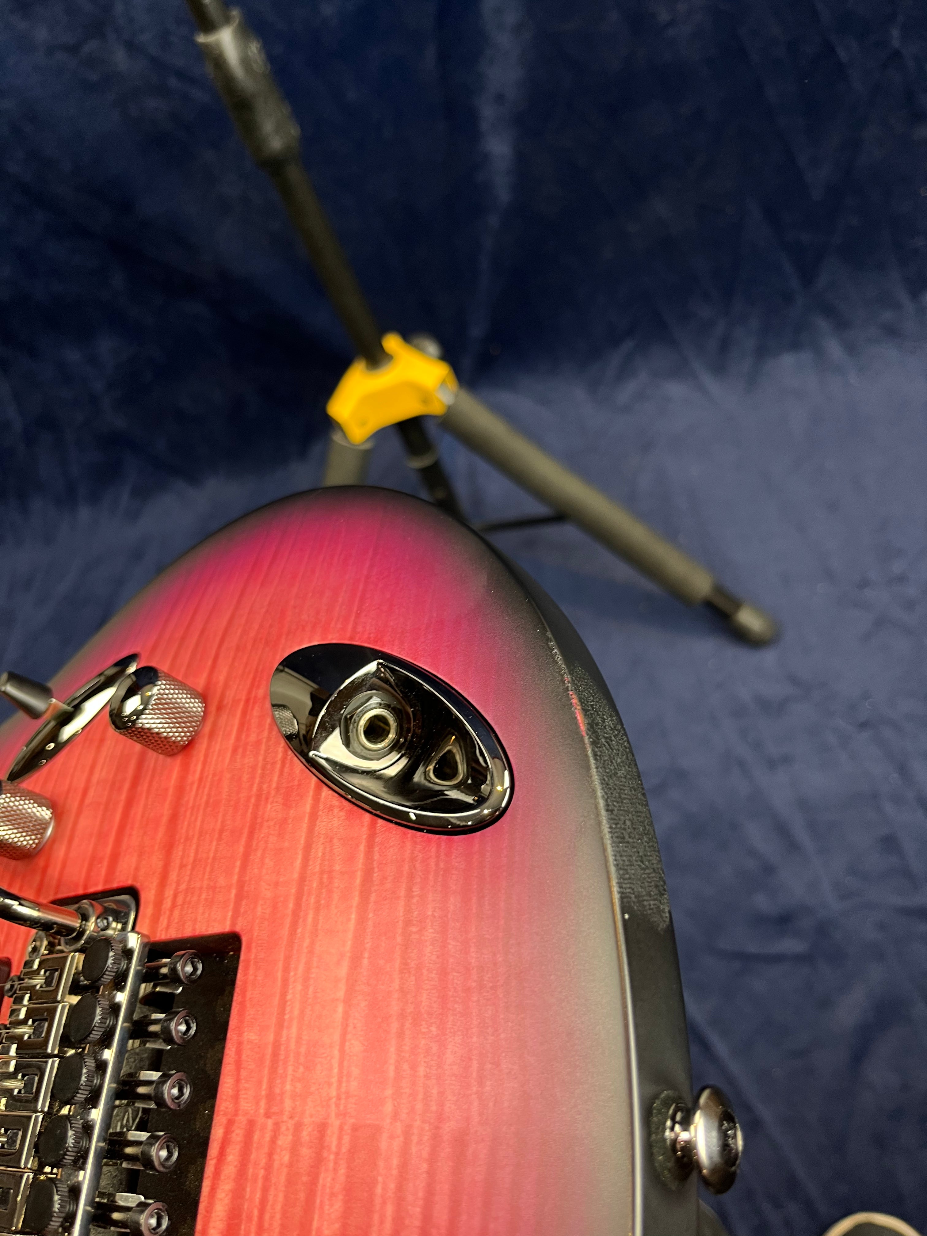 Electric guitar with a pink burst finish showing the jack plate, control knobs, and bridge, against a dark blue background with a guitar stand visible.