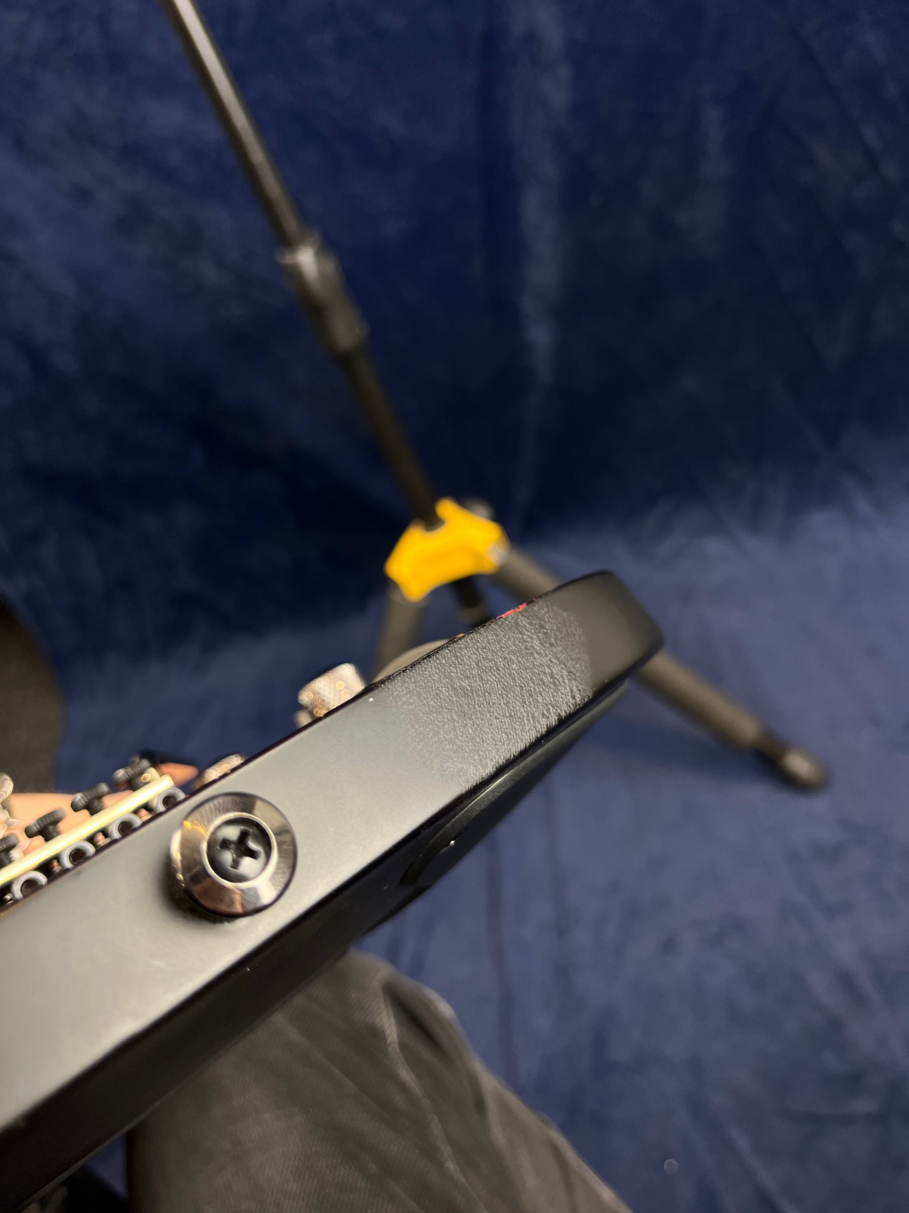 Close-up of a black electric guitar body showing a screw for the strap button and visible edge wear against a blue backdrop and tripod stand.