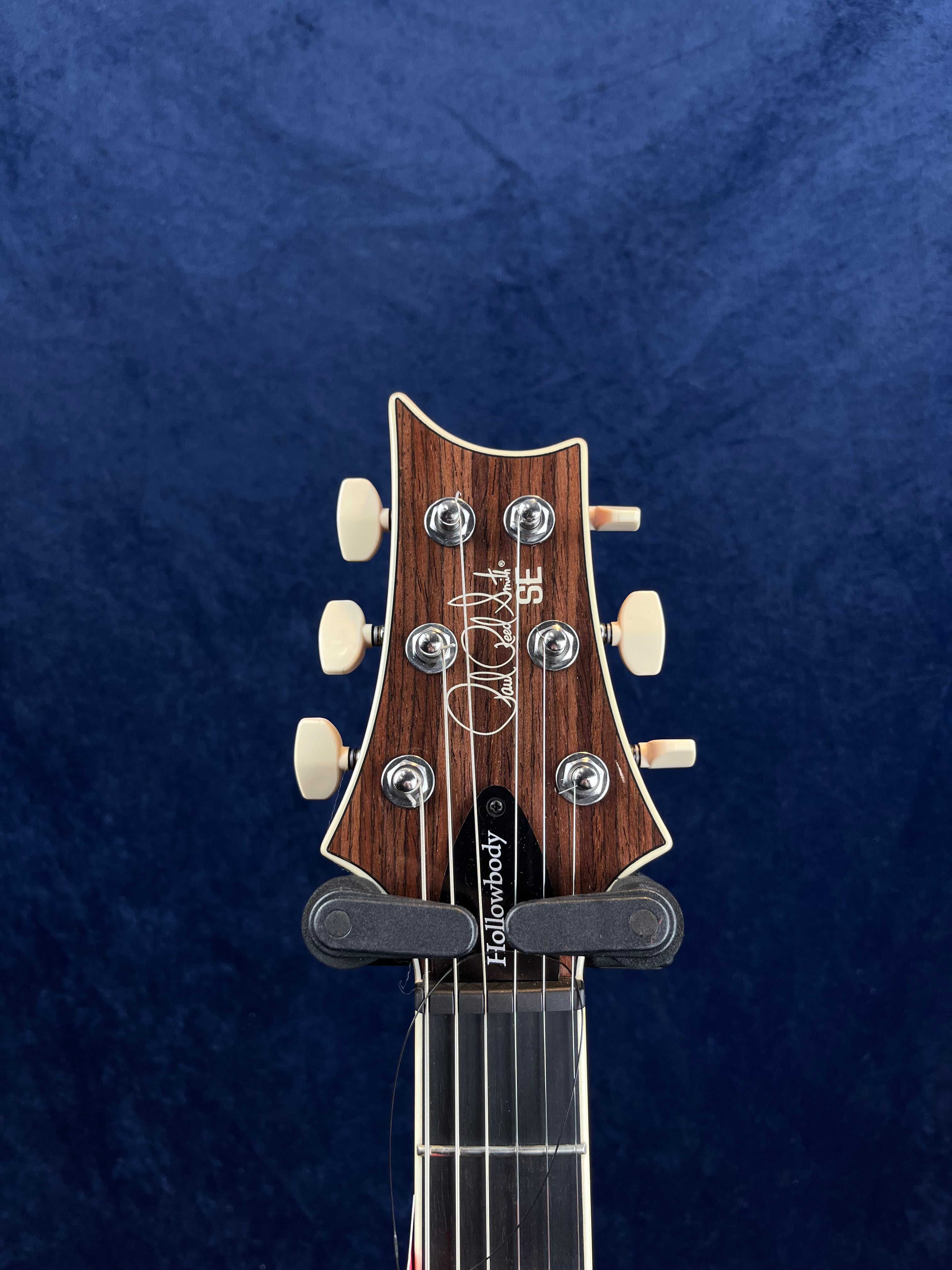 Electric guitar headstock with six vintage-style cream tuning pegs, Paul Reed Smith signature, and "Hollowbody" label against a dark blue background.