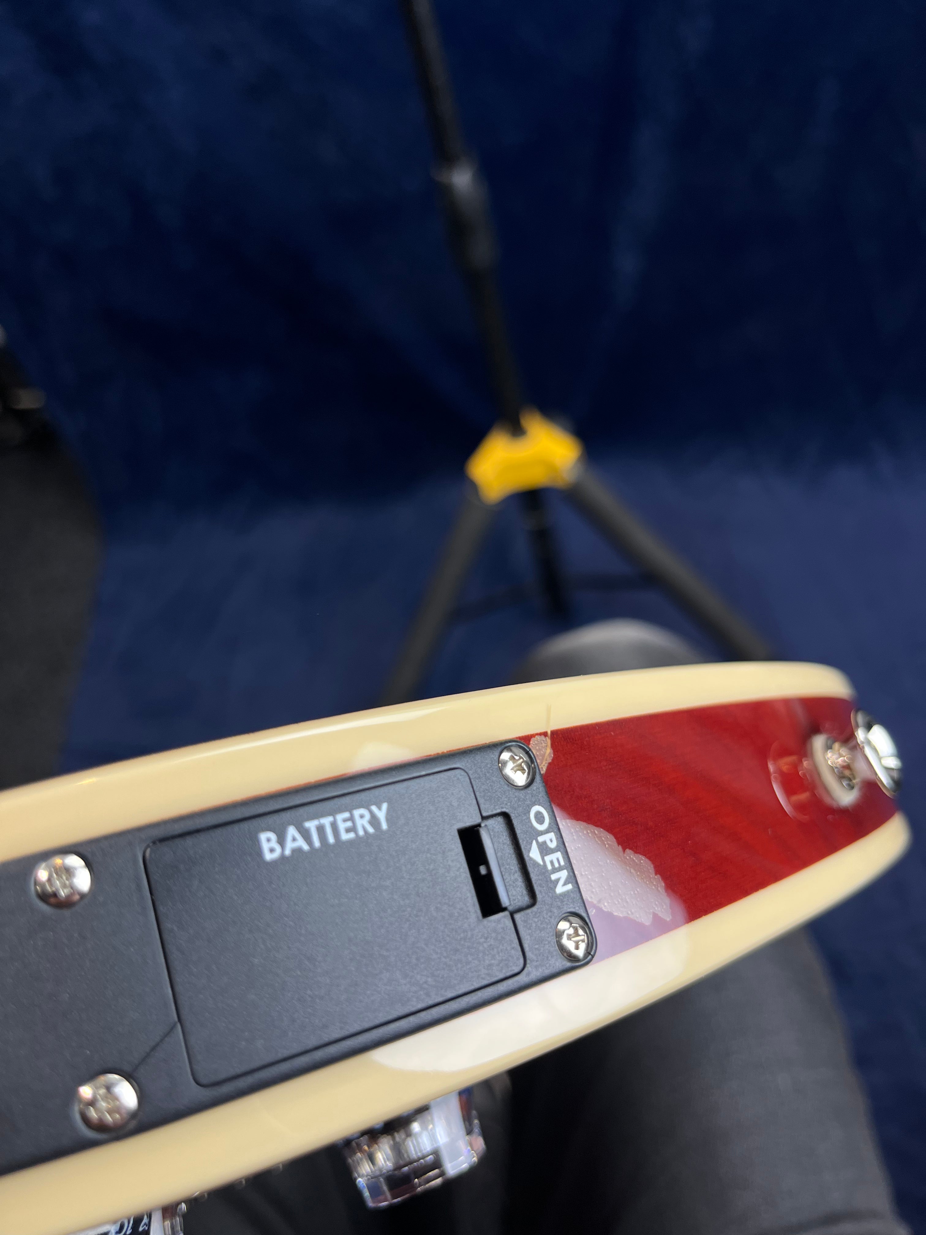 Close-up of an electric guitar body shows a battery compartment labeled "Battery" with an "Open" switch and some chipped paint on the edge.